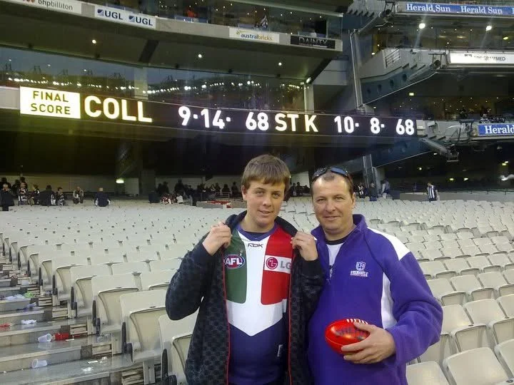 Two men standing in an empty sports stadium, smiling at the camera. One wears an AFL jersey, the other a purple jacket, and holds a red oval-shaped object. The scoreboard above shows final scores for a game date of September 14, 1968.