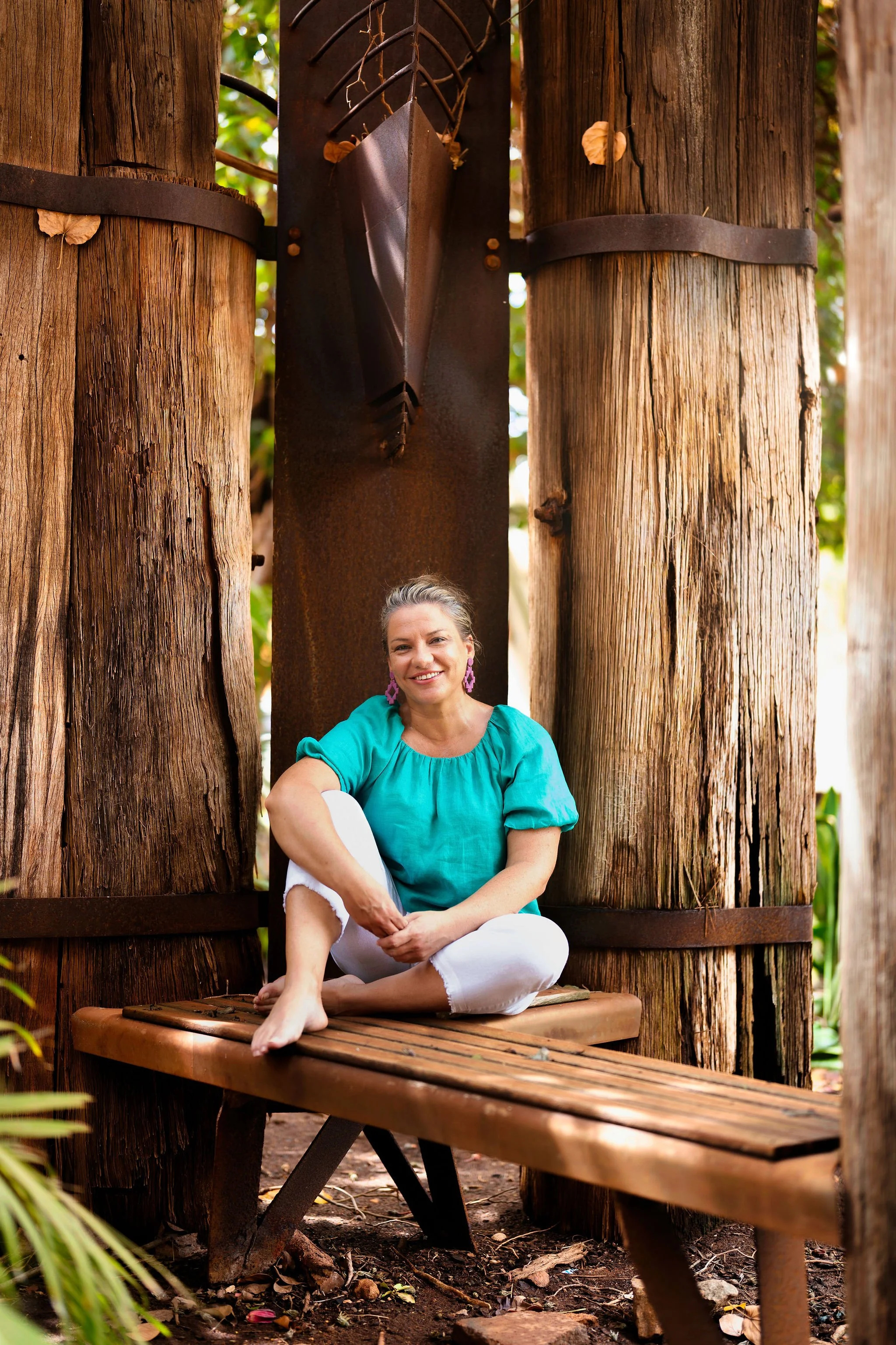 A woman sitting on a wooden bench outdoors among large wooden tree trunks, smiling, wearing a turquoise top and white pants, with pink earrings.
