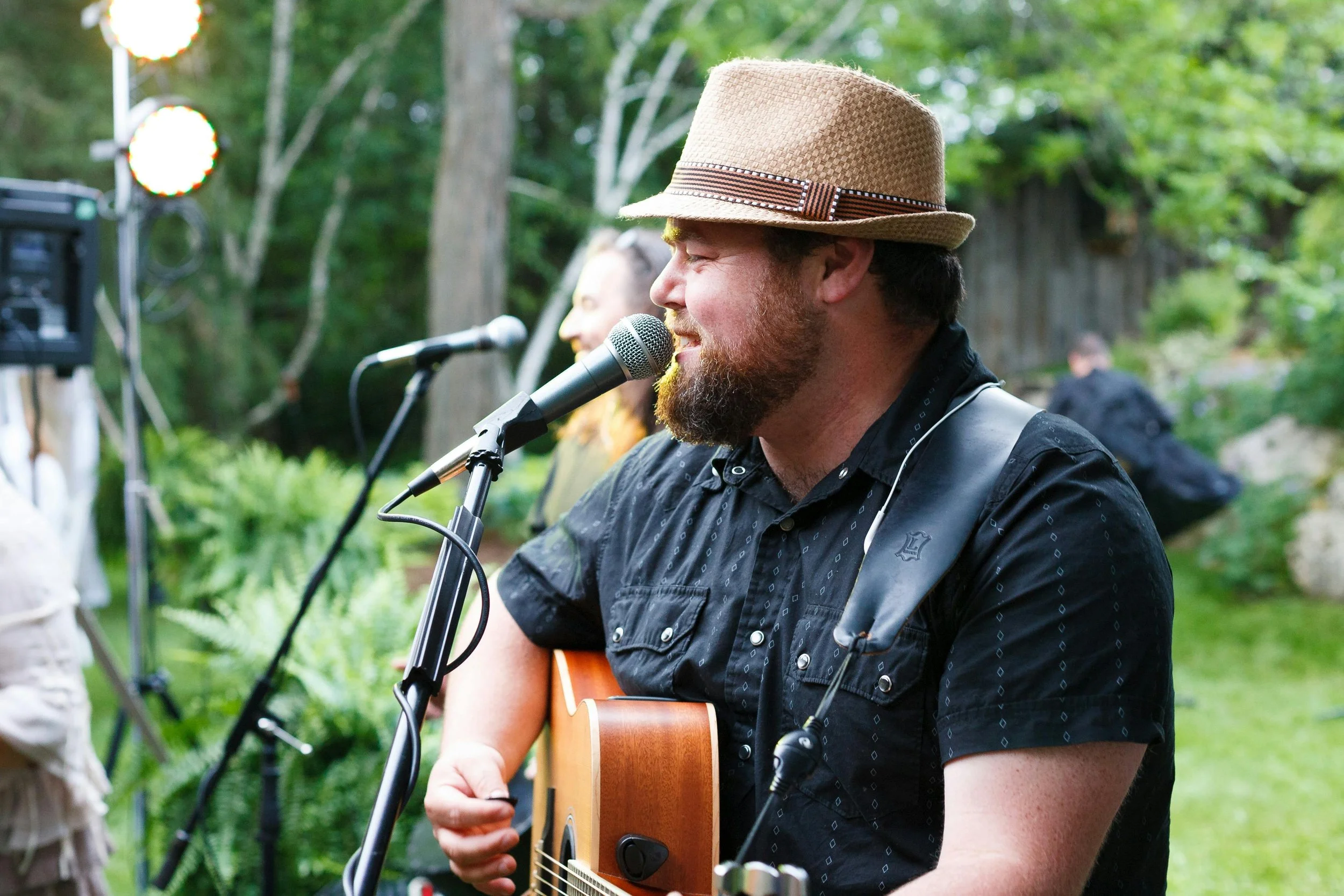 Man with a beard and a straw hat playing an acoustic guitar and singing into a microphone outdoors during daytime, with trees and other people in the background.