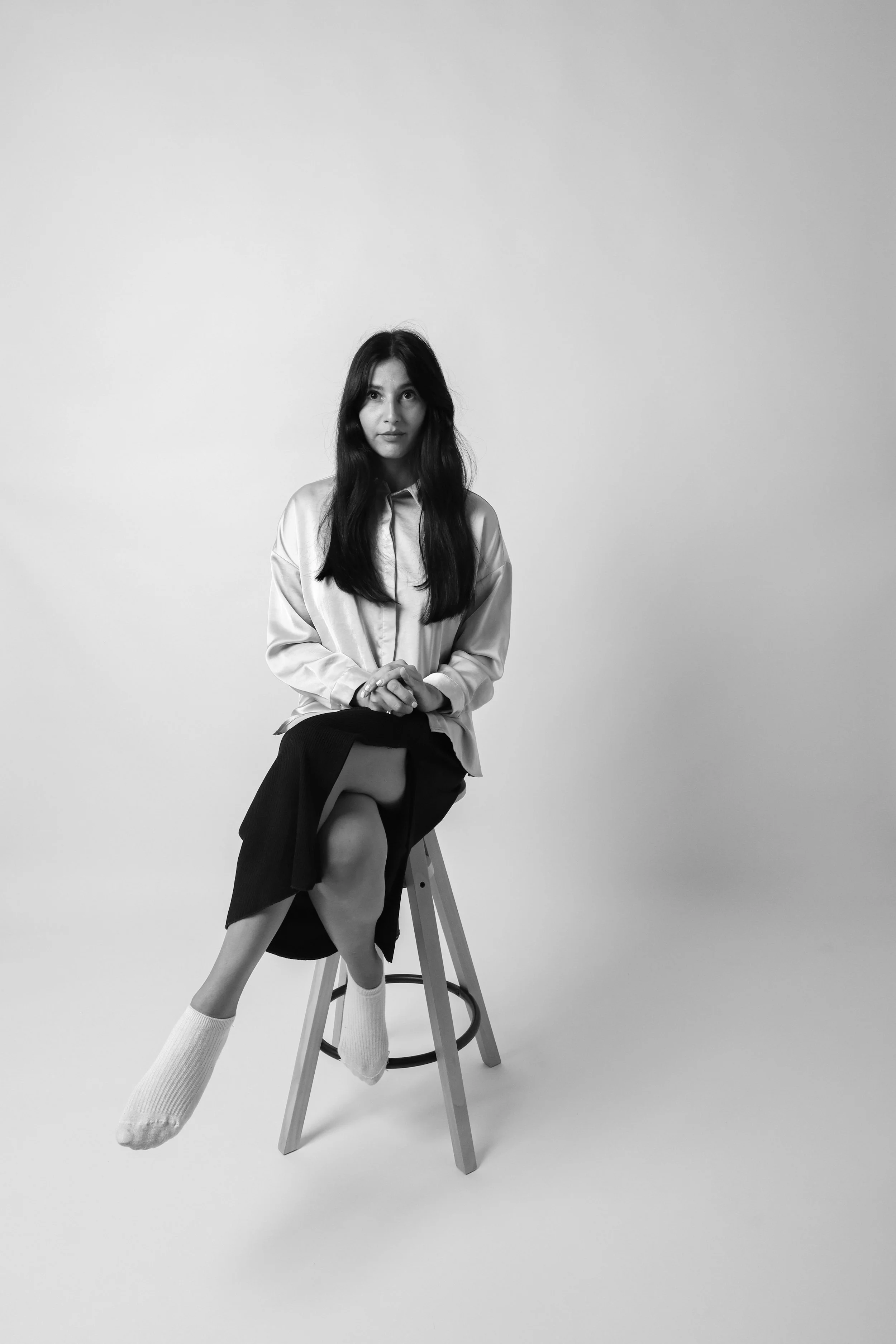Black and white photo of a woman sitting on a stool with crossed legs, wearing a light-colored shirt, a dark skirt, and white socks, posing against a plain background.