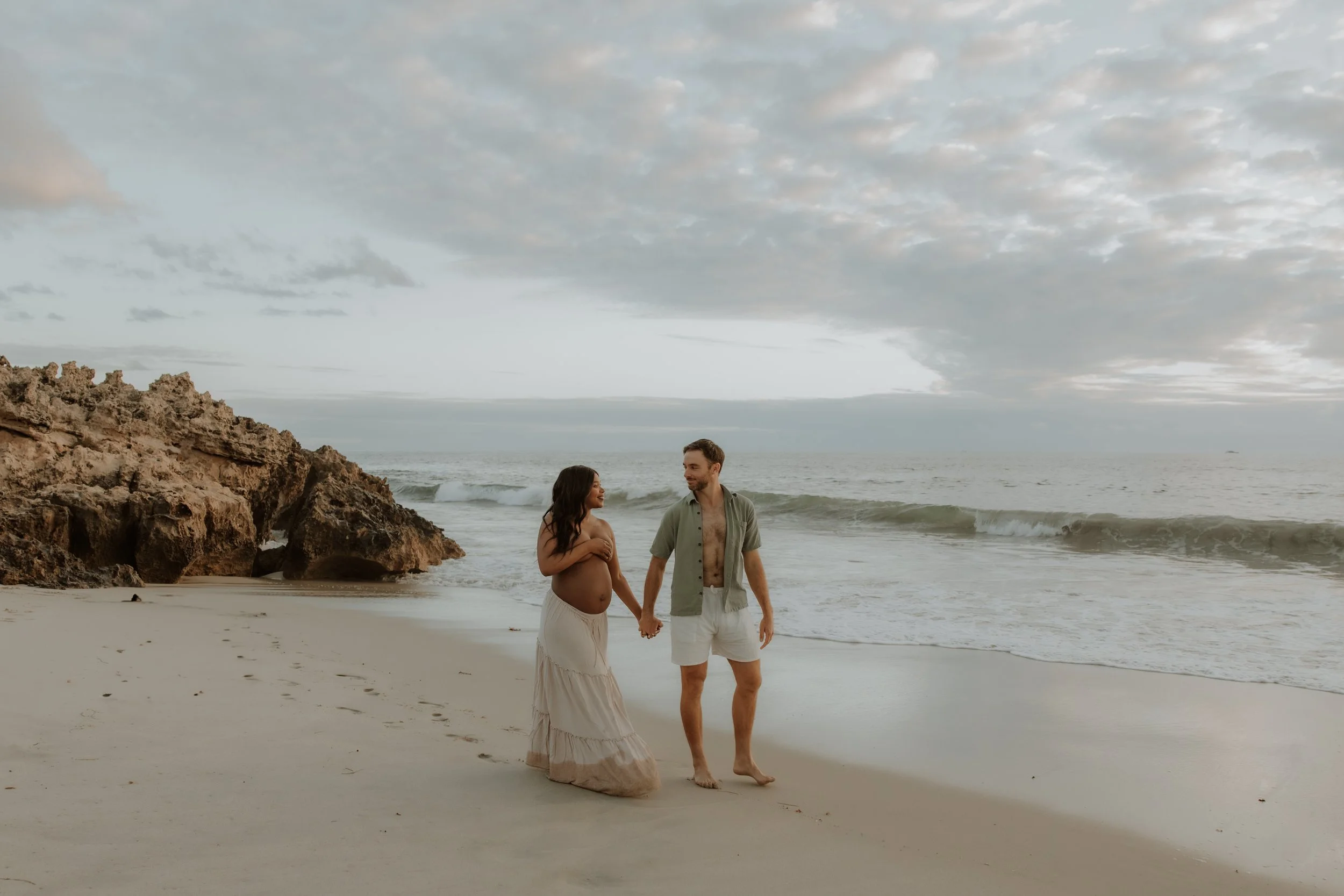 A couple holding hands on a beach, with a pregnant woman in a white skirt and a man in white shorts and an open shirt, walking near the waves at sunset.