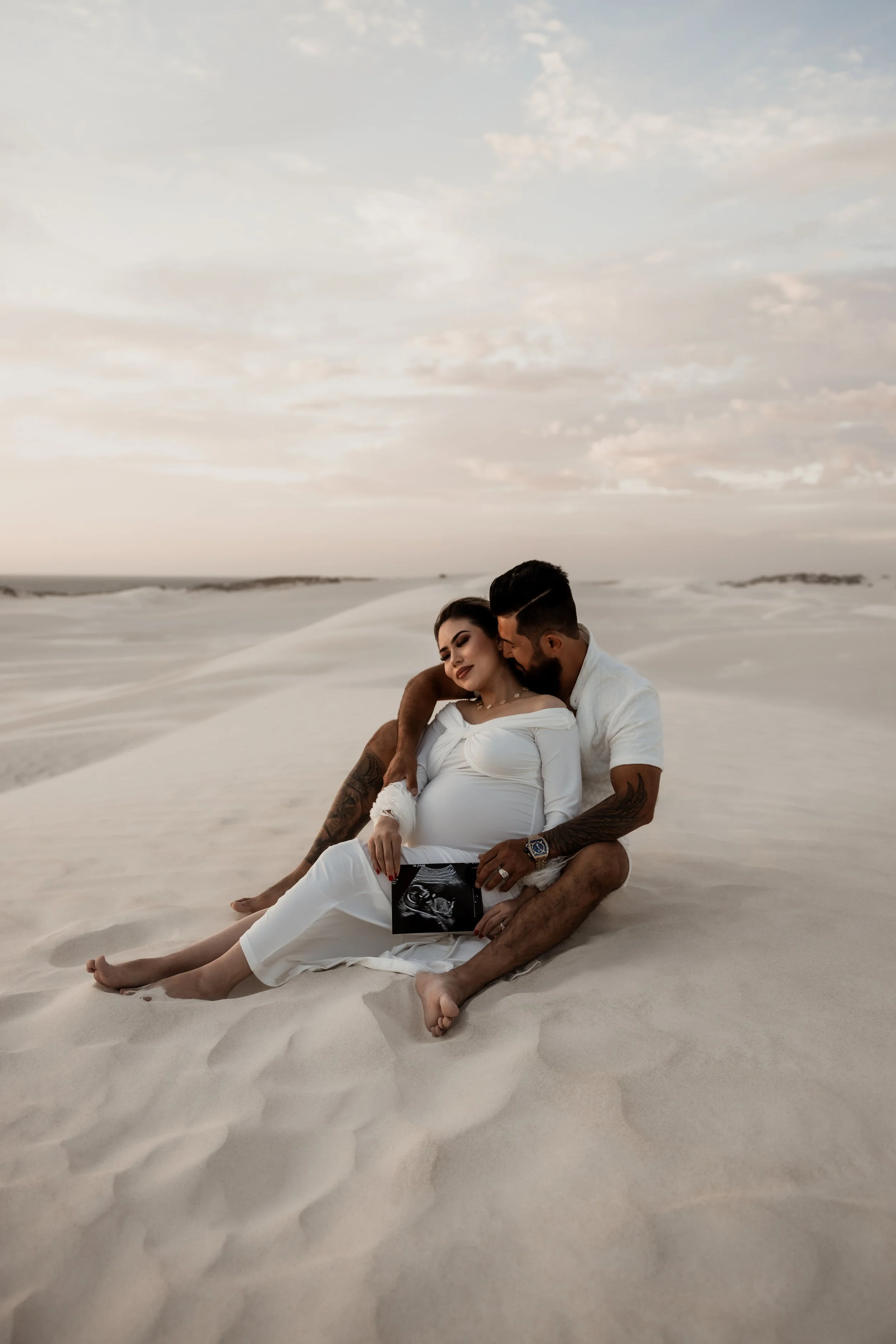 A couple sitting on sand dunes in a desert at sunset, with the woman holding an ultrasound image, indicating pregnancy. Maternity photoshoot in Lancelin.