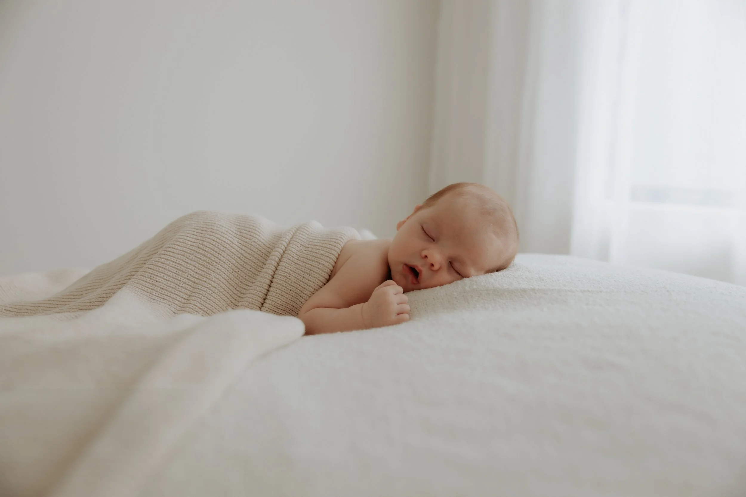 A sleeping newborn baby lying on a white bed, covered with a beige knitted blanket in a studio setting