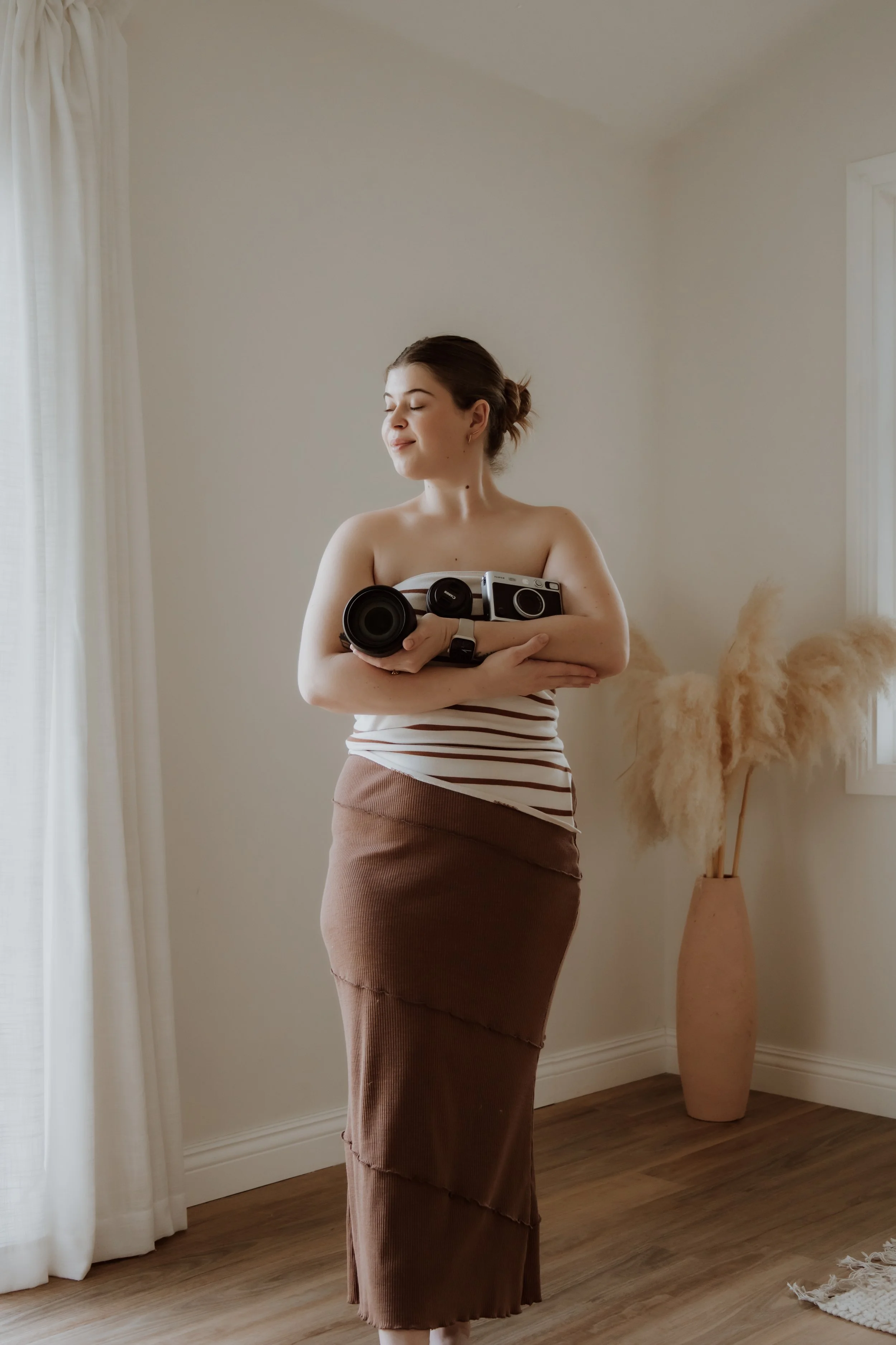 A woman with brown hair pulled back, standing in a room with light-colored walls, holding a vintage camera and a camera lens close to her chest, with eyes closed and a serene expression.