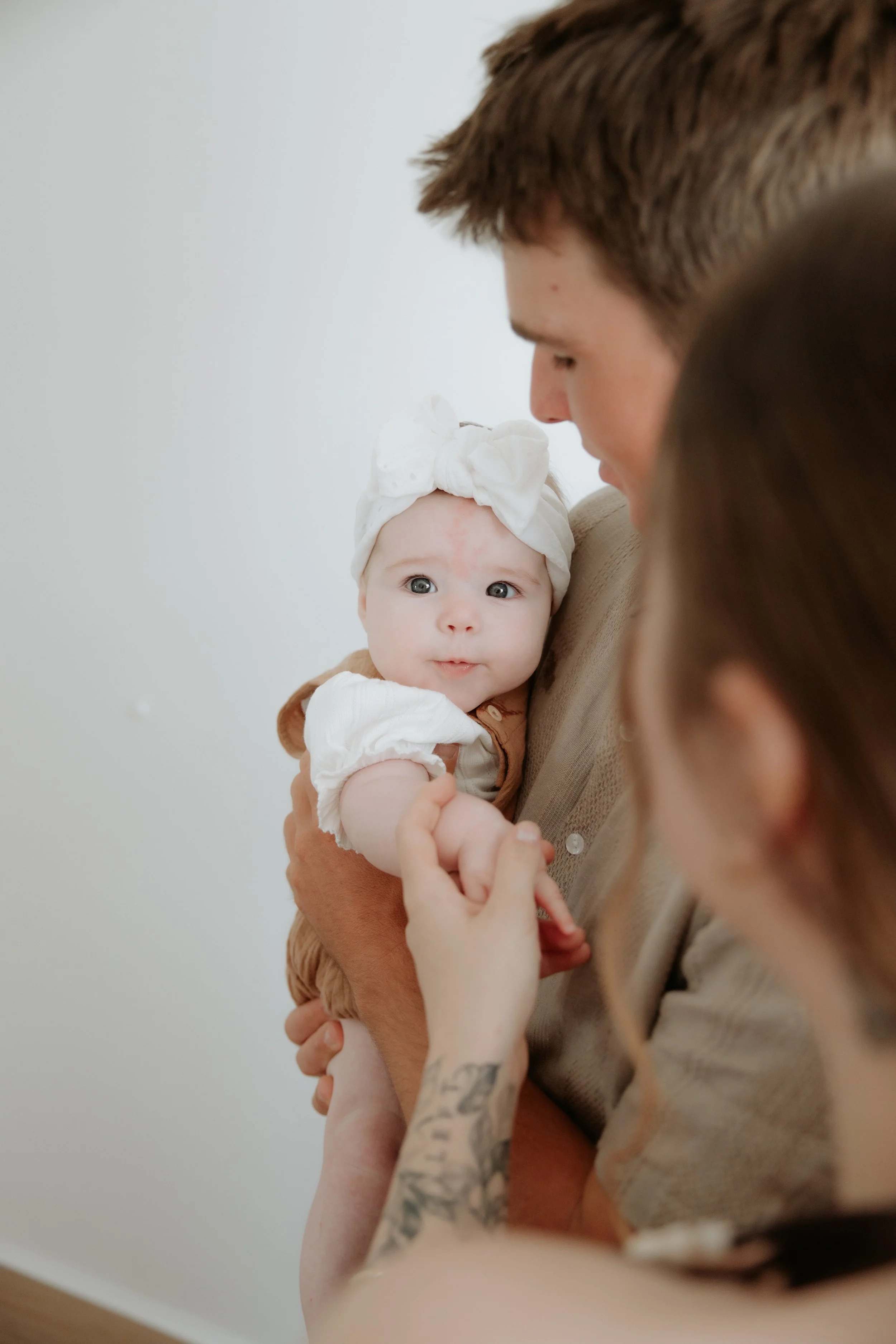 A baby girl with big blue eyes being held by a man, with a woman touching her arm, in a close-up indoor setting.