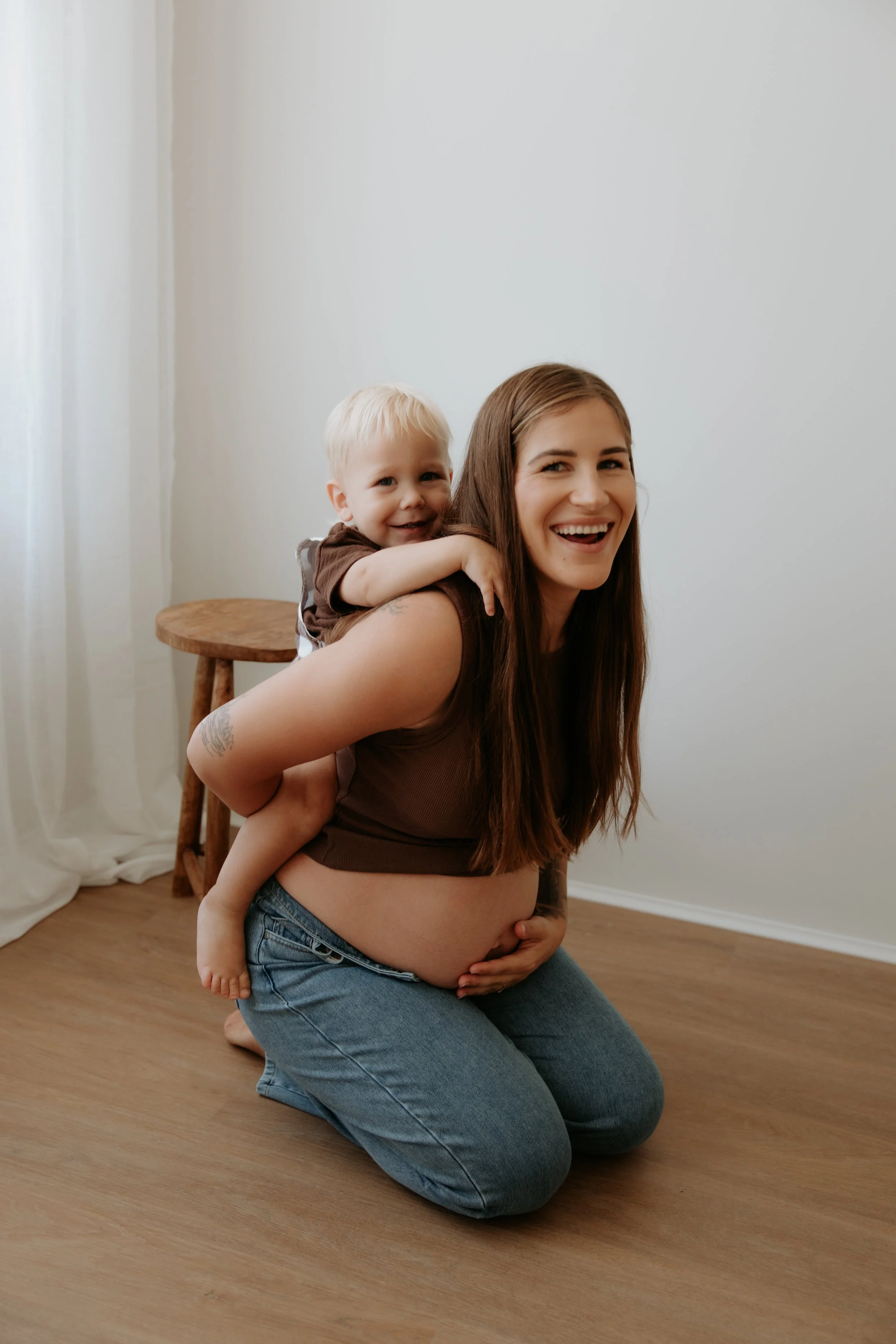 A smiling pregnant woman kneeling on the floor with a toddler on her back, in a room with light-colored walls and hardwood floor.