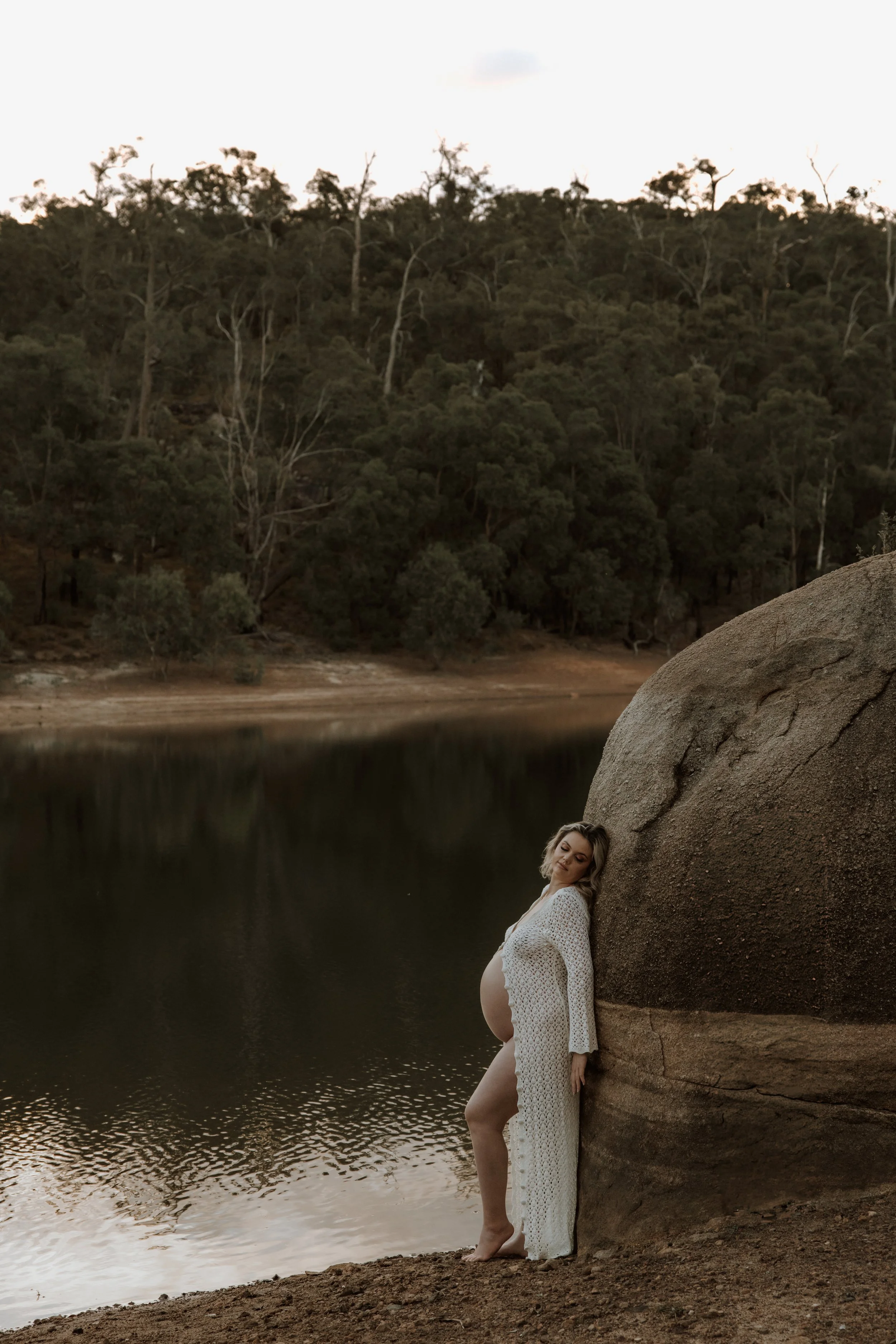 Pregnant woman in a white crochet dress standing by a large boulder next to a calm lake with a forested hillside in the background at dusk.