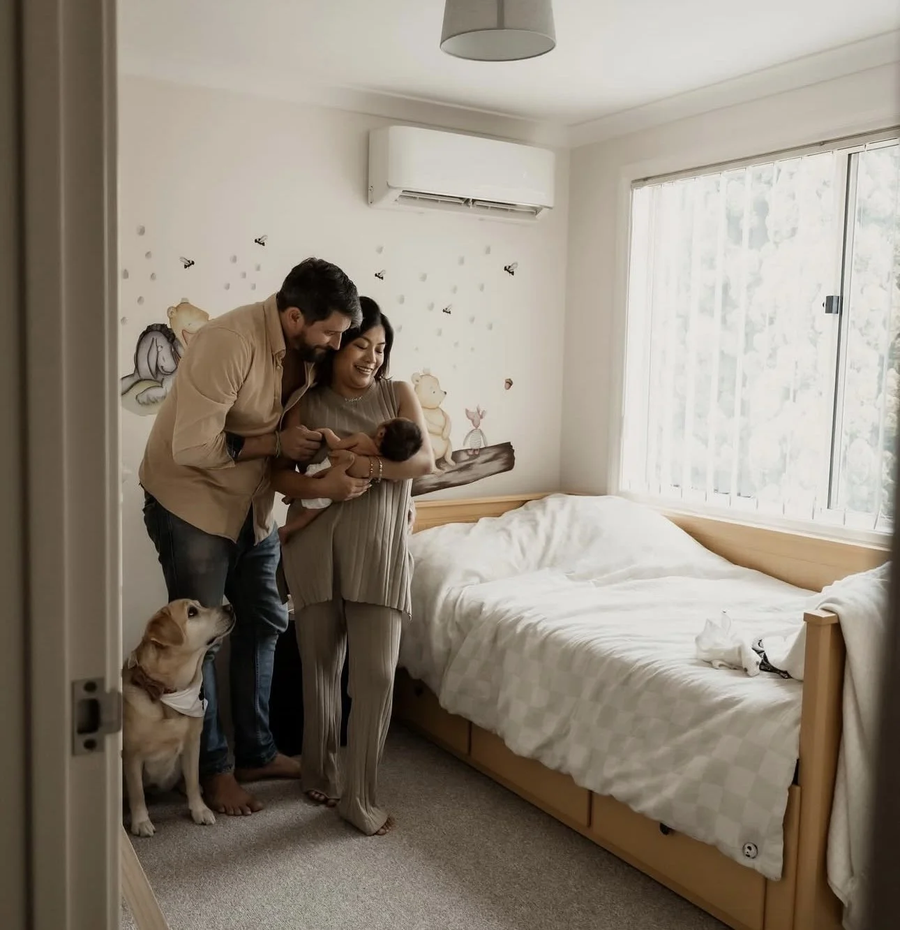 A couple holding a newborn baby in a bedroom with a dog sitting nearby. The room has white walls with butterfly and animal decals, a wooden bed with white bedding, and a large window.