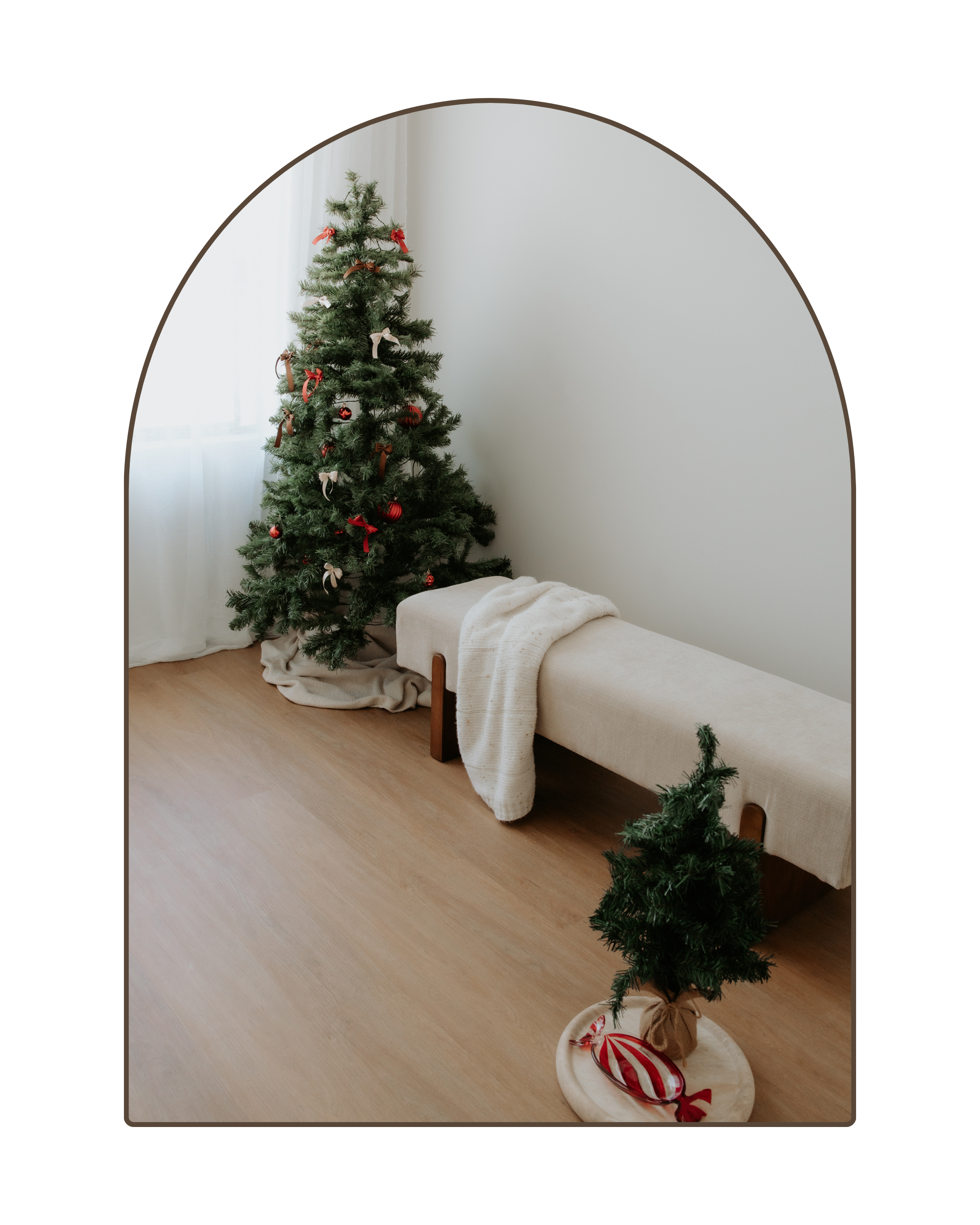 A decorated Christmas tree in a bright room with a sheer white curtain. A small artificial tree is on the floor on a round wooden tray, decorated with candy and ribbon. A beige bench with a white blanket is beside the tree and a larger tree with red and white ornaments and ribbons is in the background.