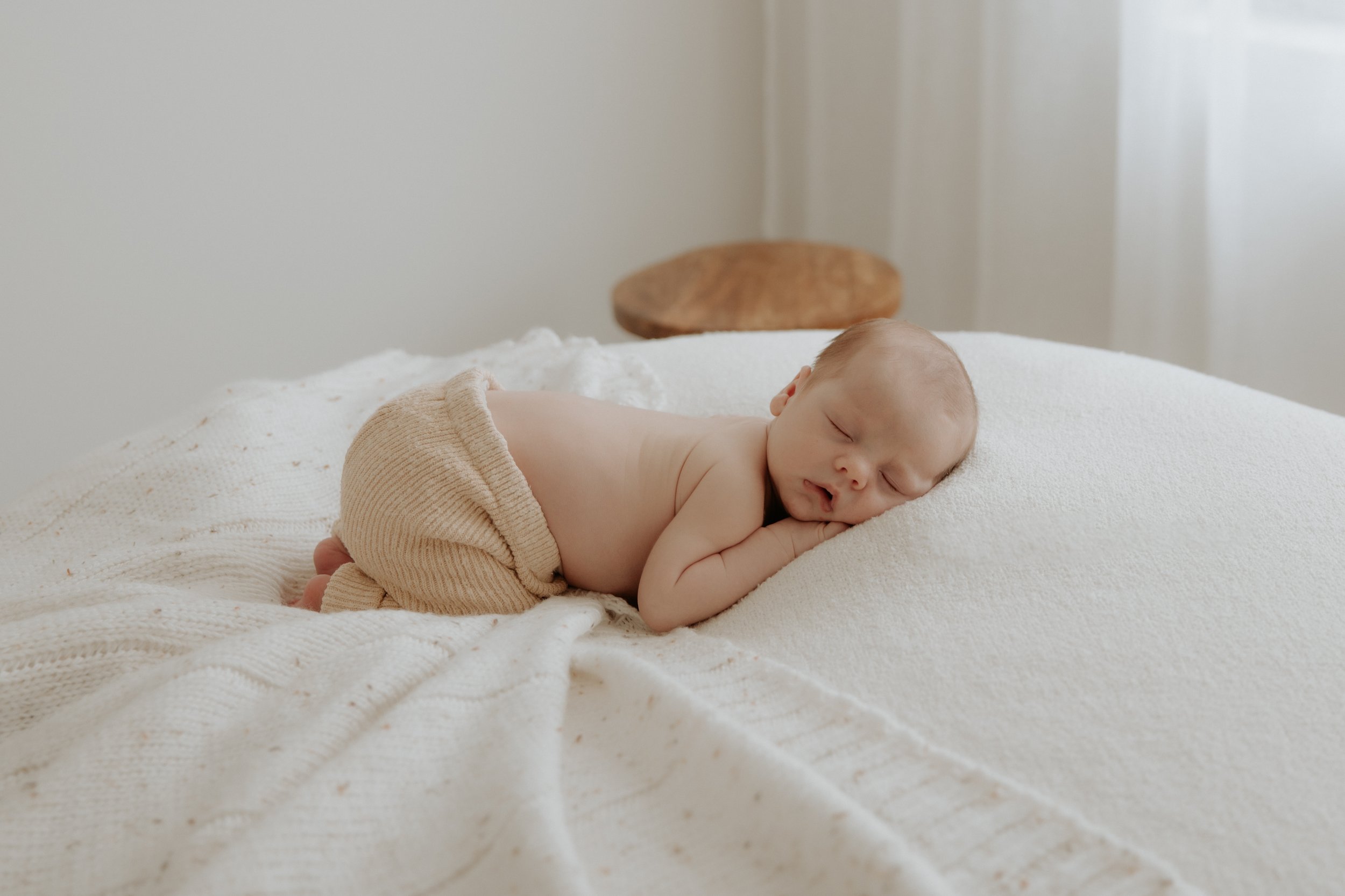A newborn baby sleeping on a white textured blanket on a bed, wearing beige knitted pants. Simple baby boy in minimal and true to colour studio photoshoot