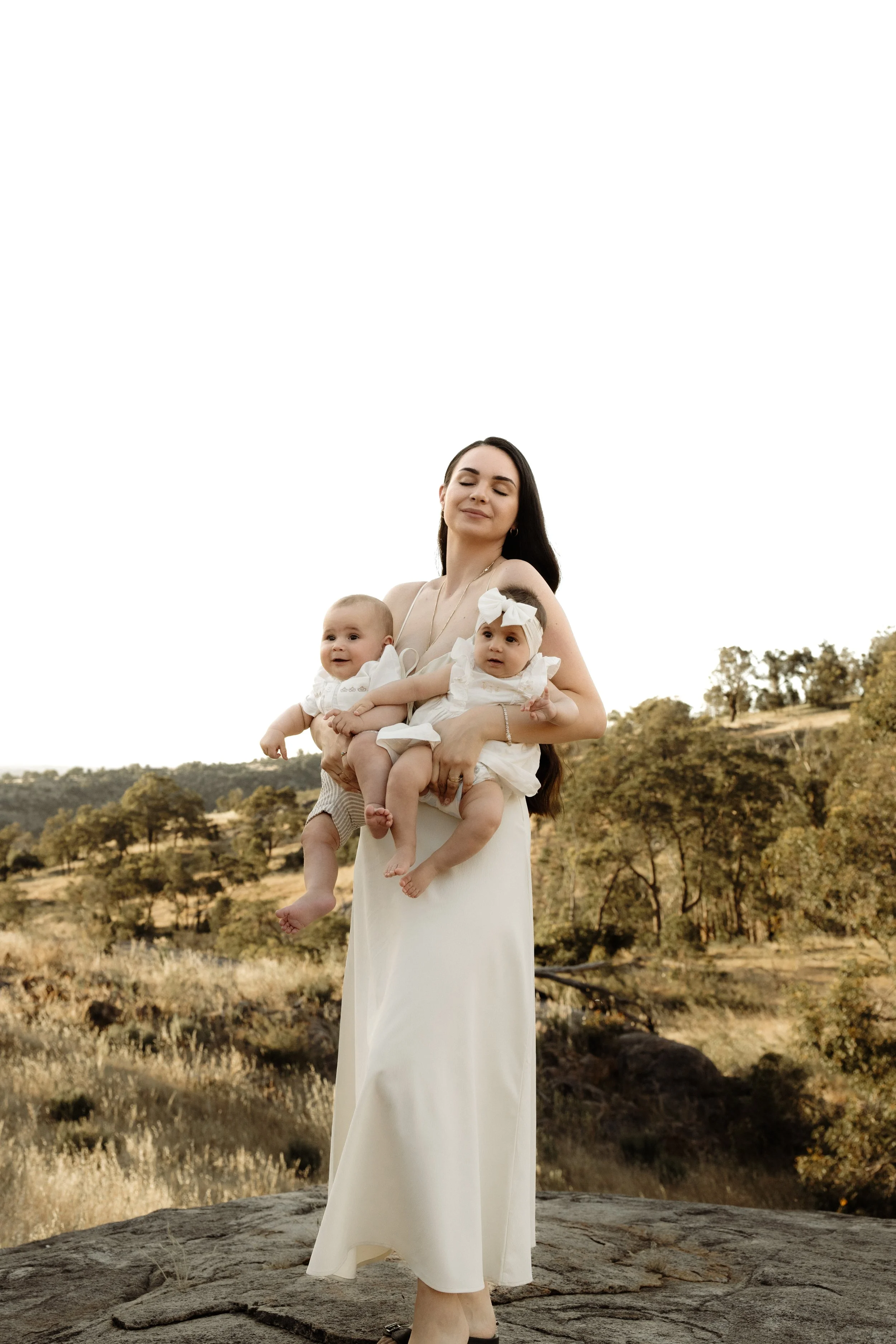 A woman in a white dress holds two babies outdoors on a rocky surface with grassy hills and trees in the background.
