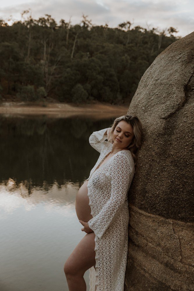 Pregnant woman leaning against a large rock by a calm lake with a forested hillside in the background, during sunset or early evening for maternity photoshoot inspiration