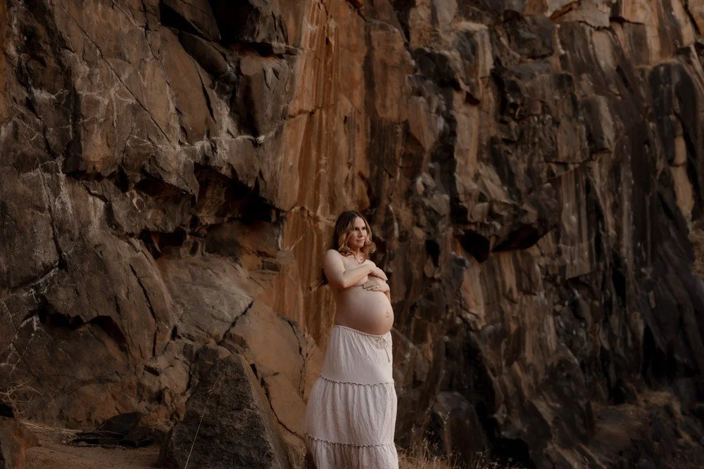Pregnant woman standing against a rocky cliff face, wearing a white skirt and topless, with arms crossed over her stomach. Unique, editorial,magazine style maternity photoshoot perth.