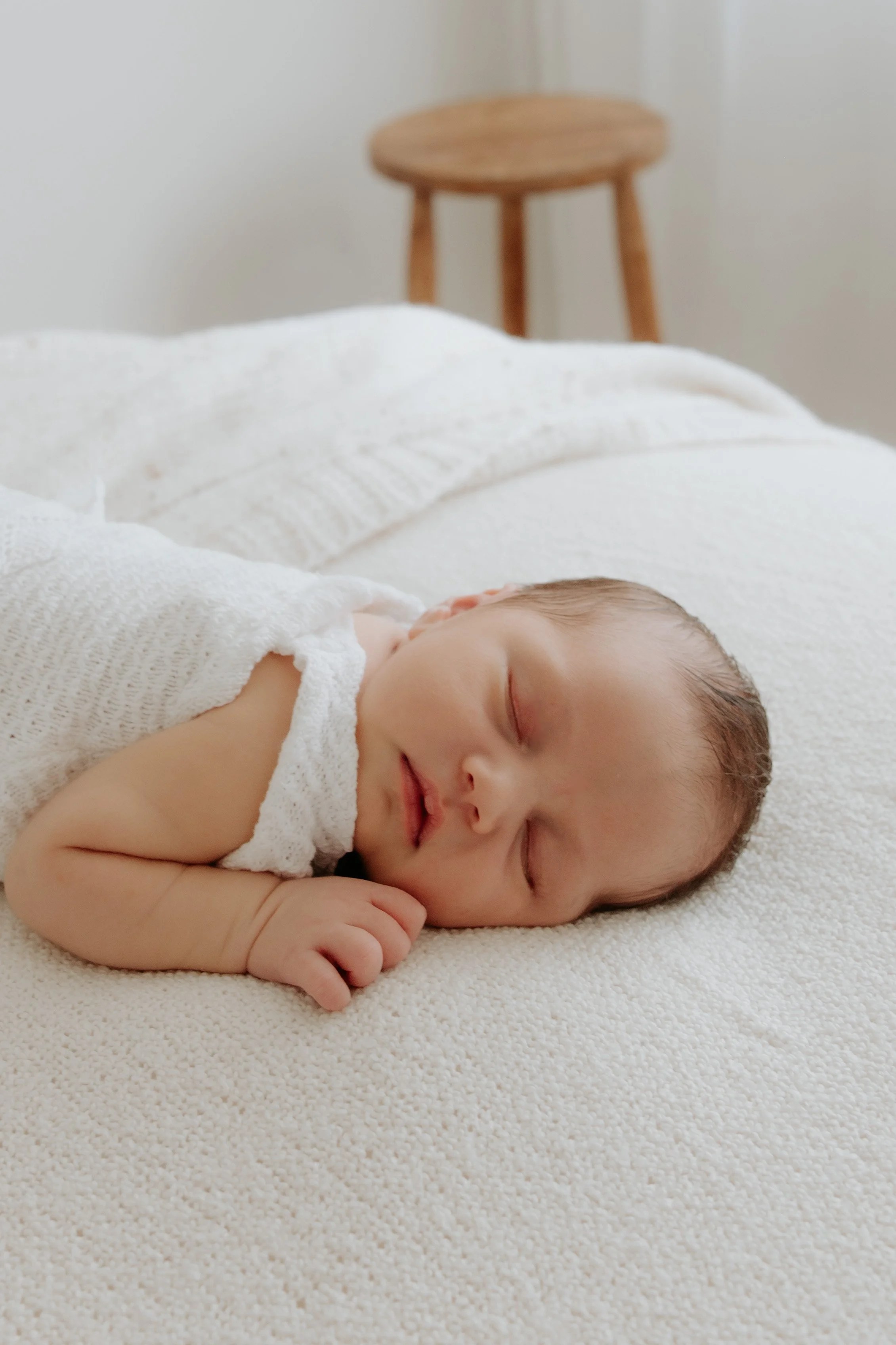A sleeping newborn baby lying on a white textured surface with a small smile, wearing a white knitted outfit. Happy newborn studio session. In the background, there is a wooden stool against a plain white wall for a minimal studio setting.
