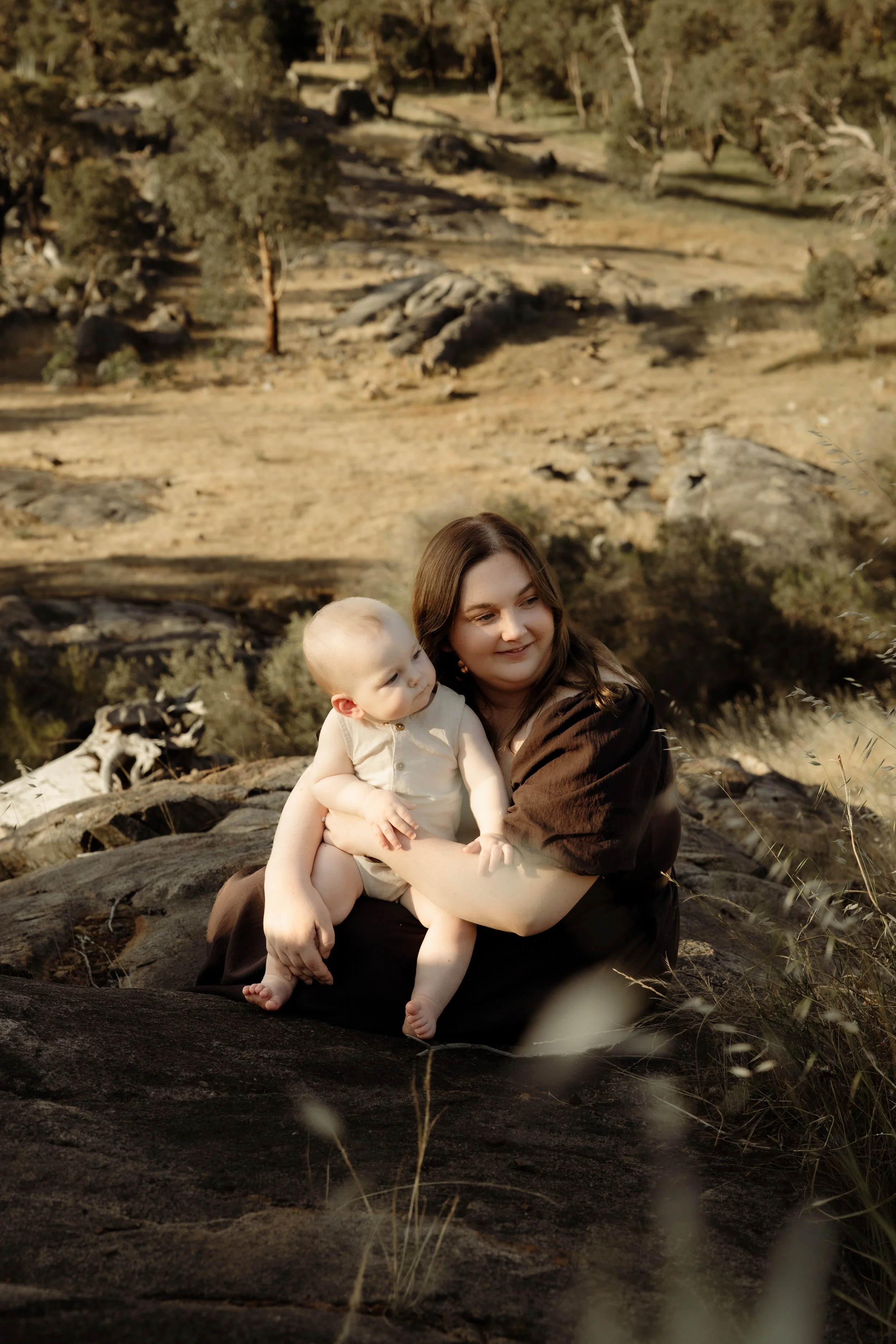 A woman holding a baby outdoors on a large rock with a natural landscape of trees and rocky terrain in the background.
