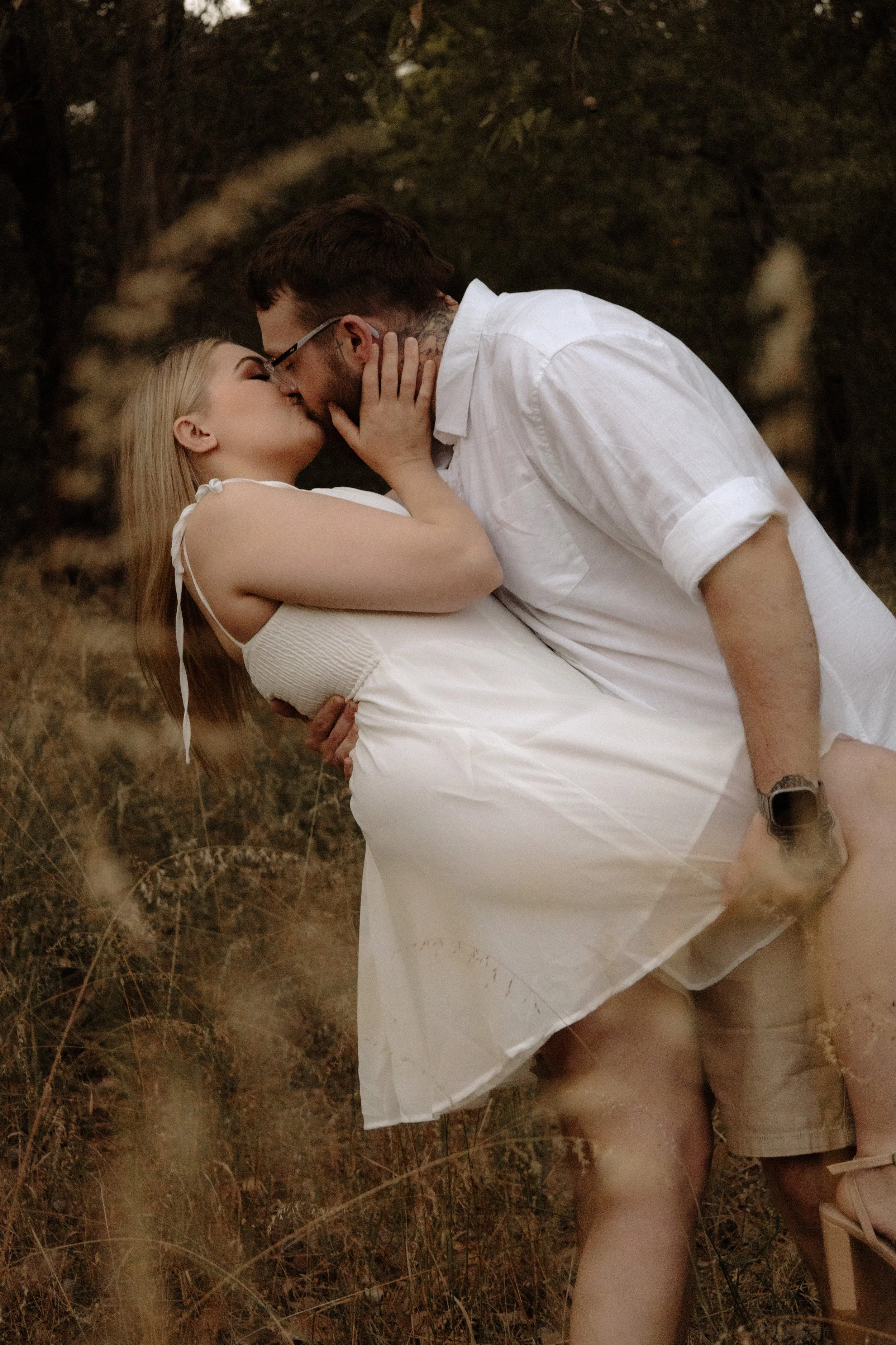 A couple is kissing in a natural outdoor setting, with the woman kneeling and the man leaning over her, both dressed in white.