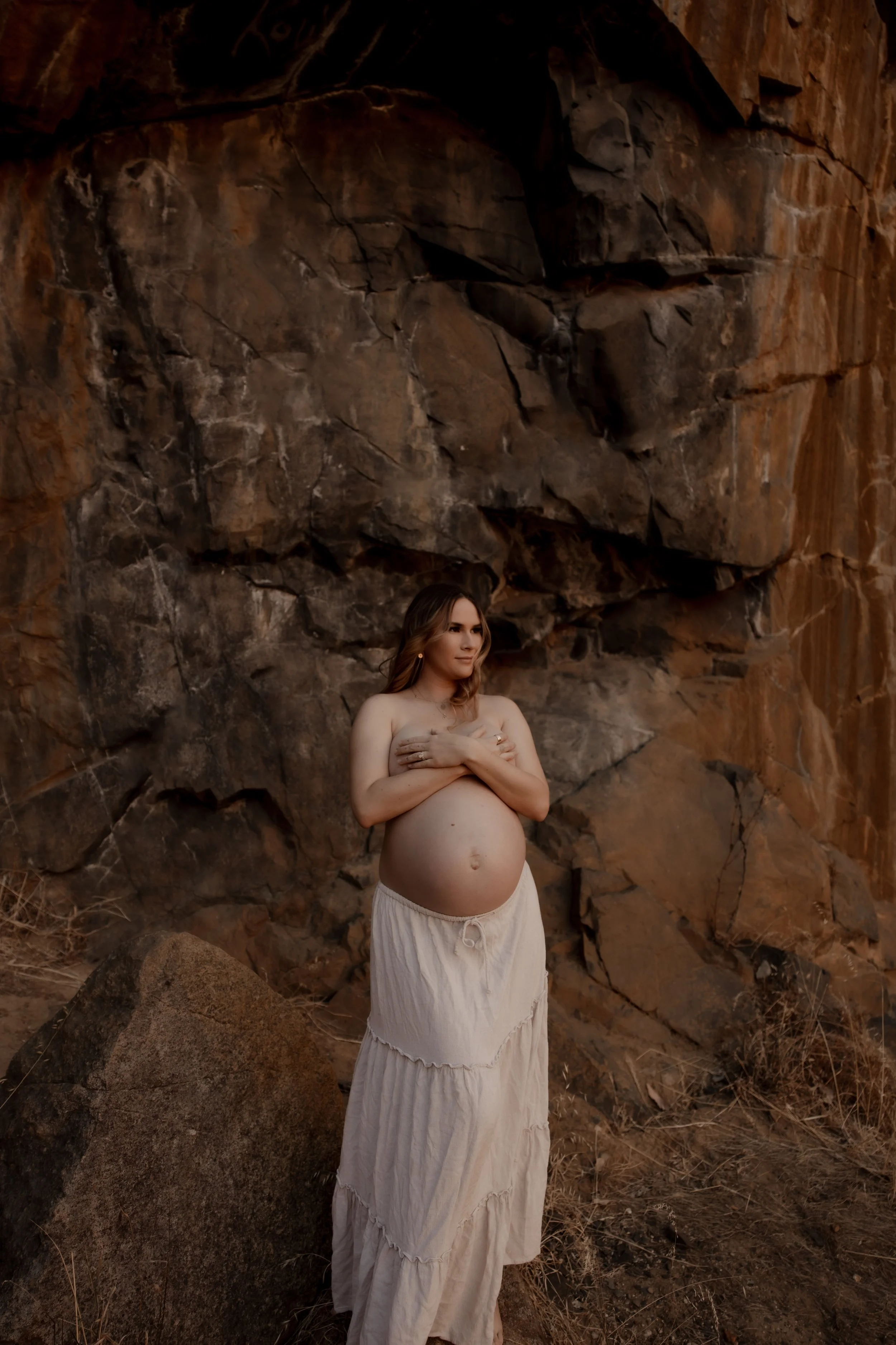 A pregnant woman standing outdoors in front of a rocky cliff, wearing a long beige skirt and holding her arms over her chest.