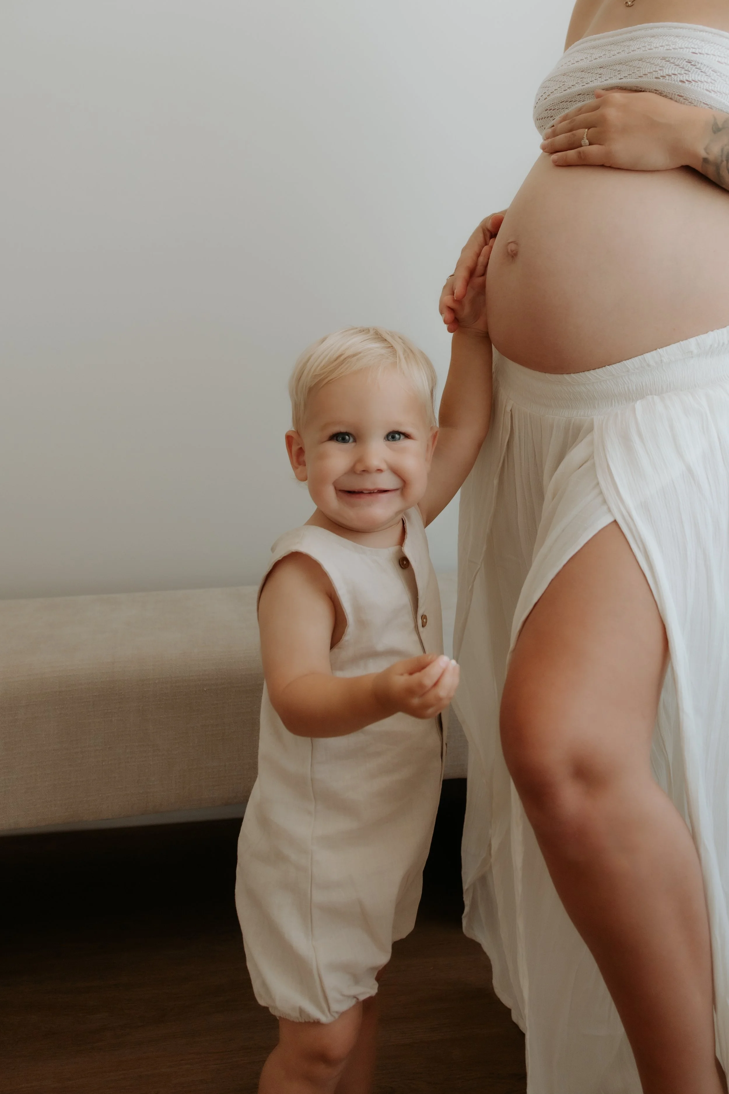 A smiling young boy touching a pregnant woman's belly, standing indoors near a beige sofa.