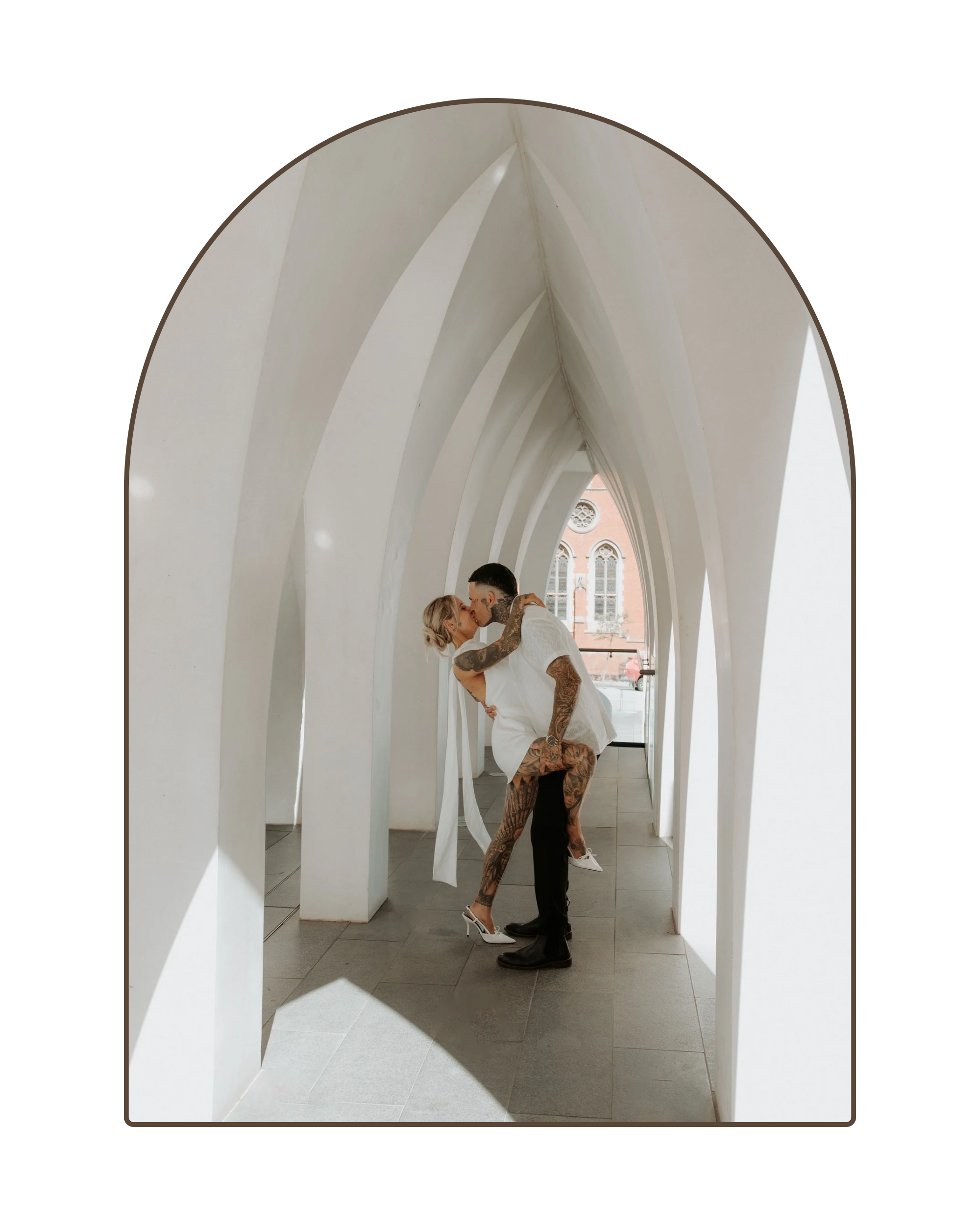 A couple sharing a kiss inside a white, modern architectural structure with arched openings, and a building visible in the background.