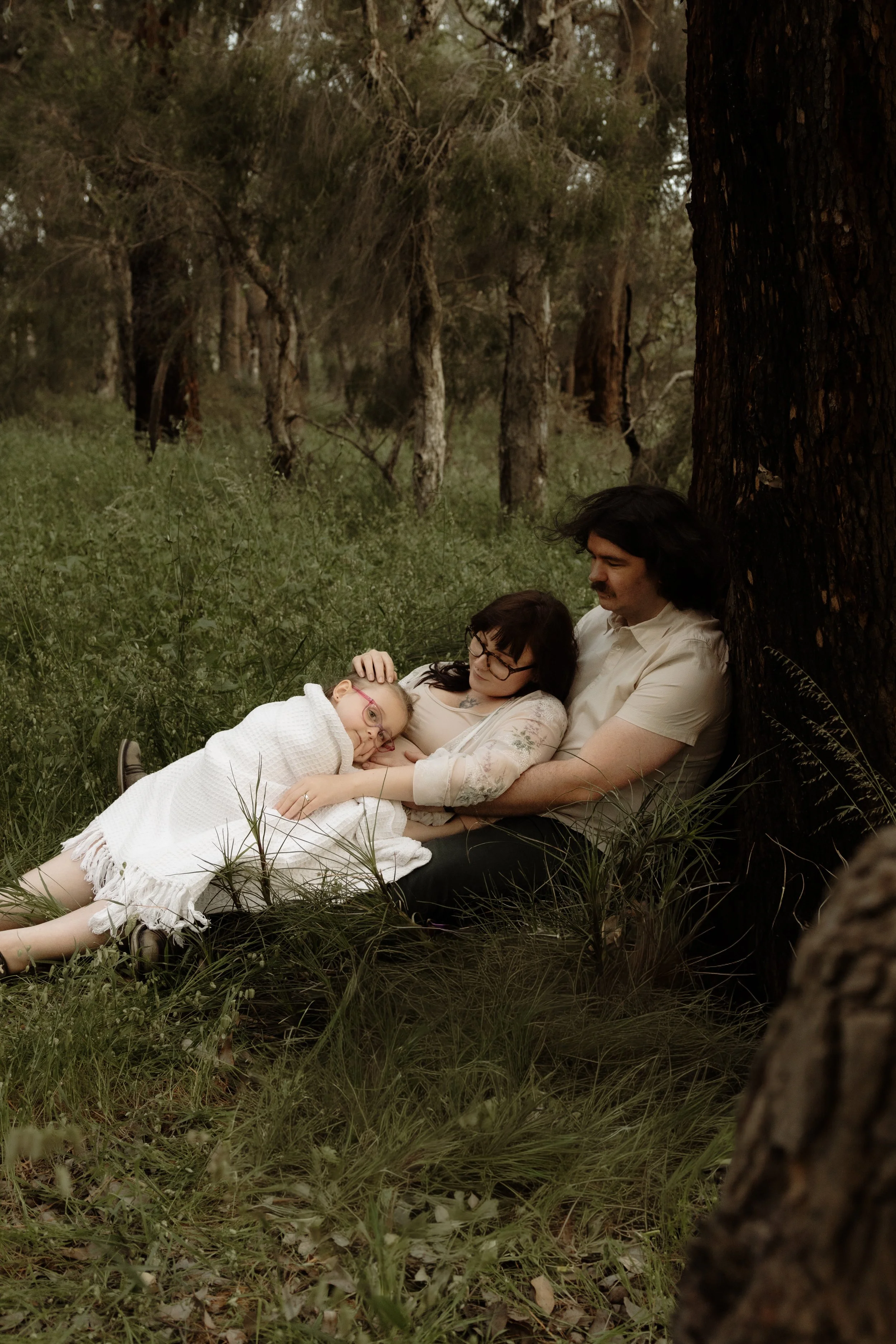 A family of three sitting on the ground in a forest, leaning against a large tree. The mother and child are cuddling, with the child resting her head on the mother's lap. The father sits nearby, looking at the mother and child. The scene is peaceful with green grass and trees around them.