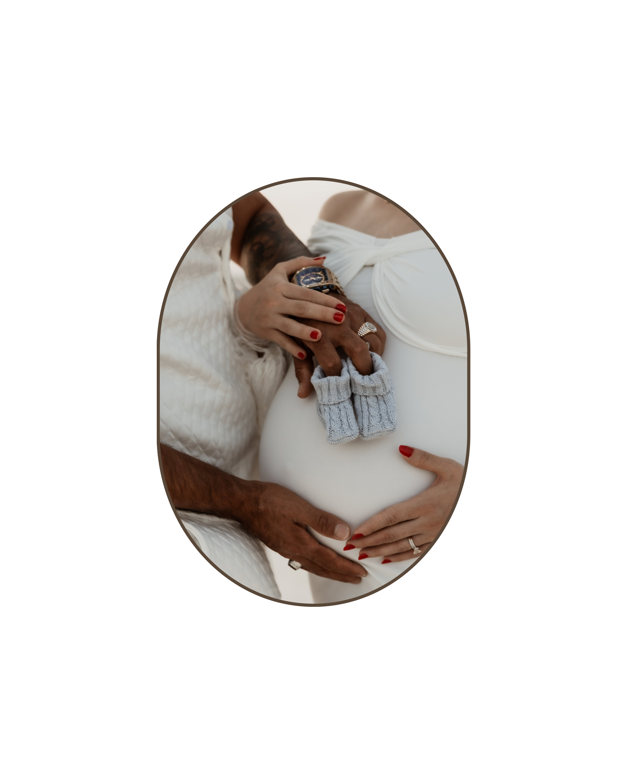 A couple's hands, one with a wedding ring, gently cradle a pregnant woman's belly, with a small baby sock and glove resting on her stomach.