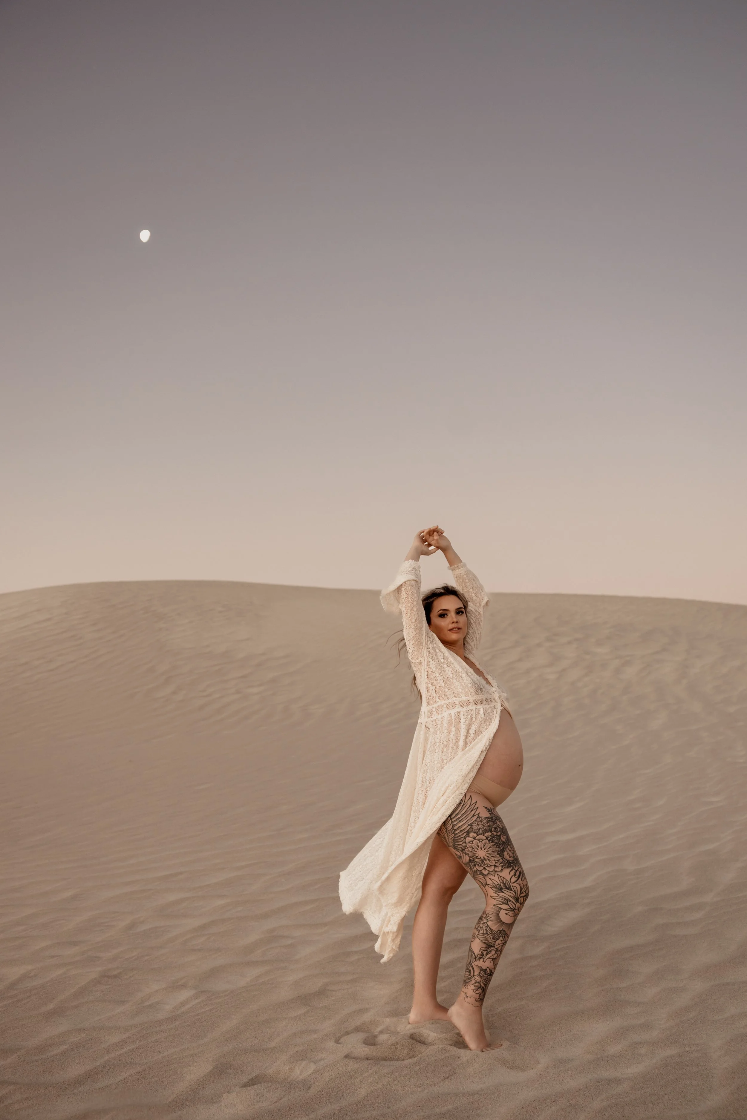 Pregnant woman standing barefoot in desert sand dunes during dusk, wearing a flowing white lace dress, with her arms raised above her head and a moon visible in the sky. Ethereal, beautiful Lancelin maternity sand dunes photoshoot Perth, WA.