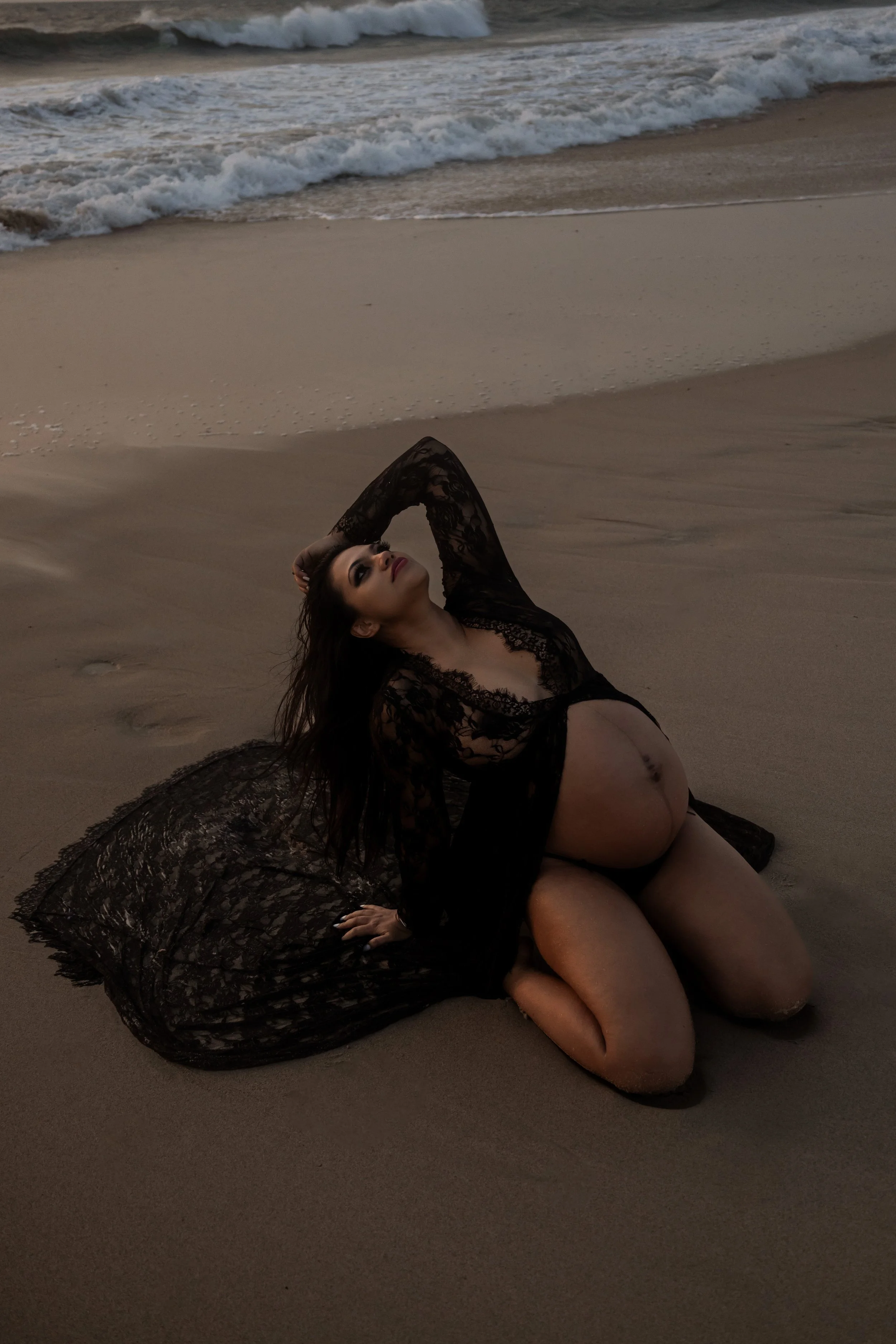 A woman in a black lace dress kneeling on the sand at the beach near the ocean, with waves in the background and overcast lighting.