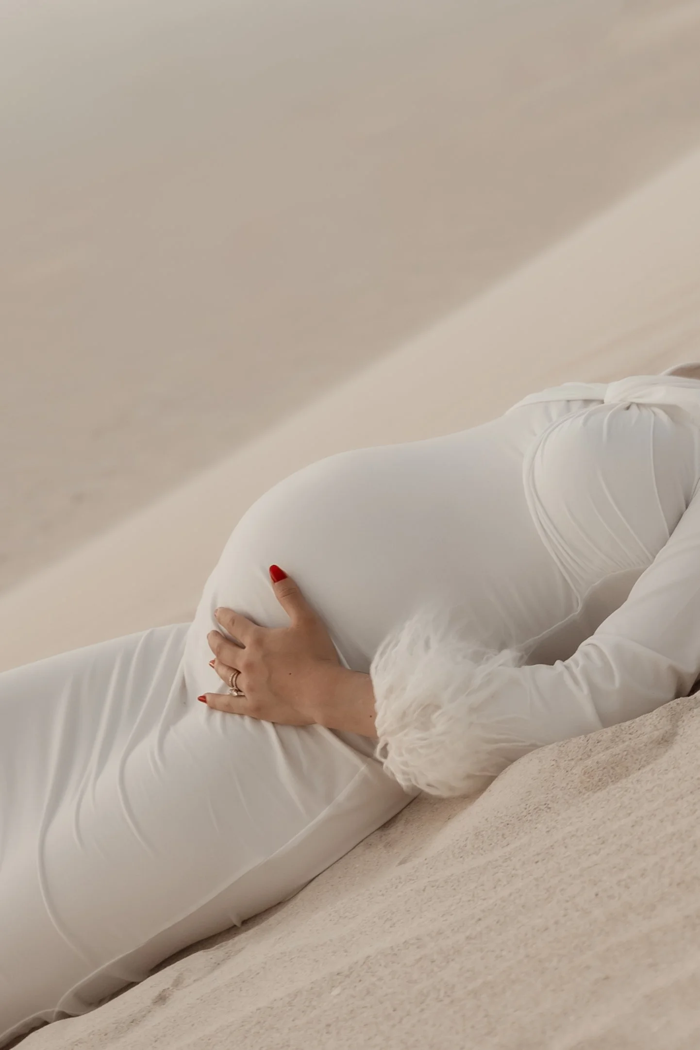 A person in a long white dress lying on their side on a beige carpeted floor, with their hand resting on their pregnant belly, showing red nail polish and a ring.