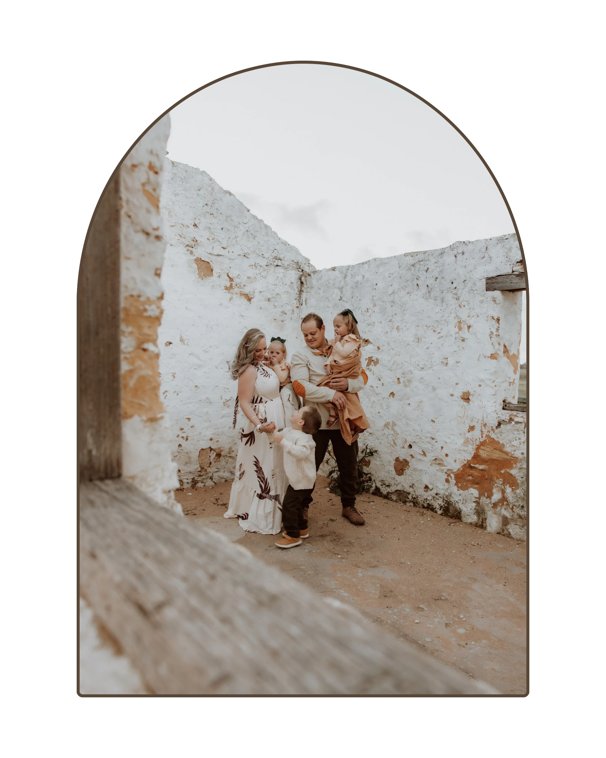 A family of five standing inside a partially ruined white-washed stone building, seen through an arched window. They are smiling and interacting with each other.