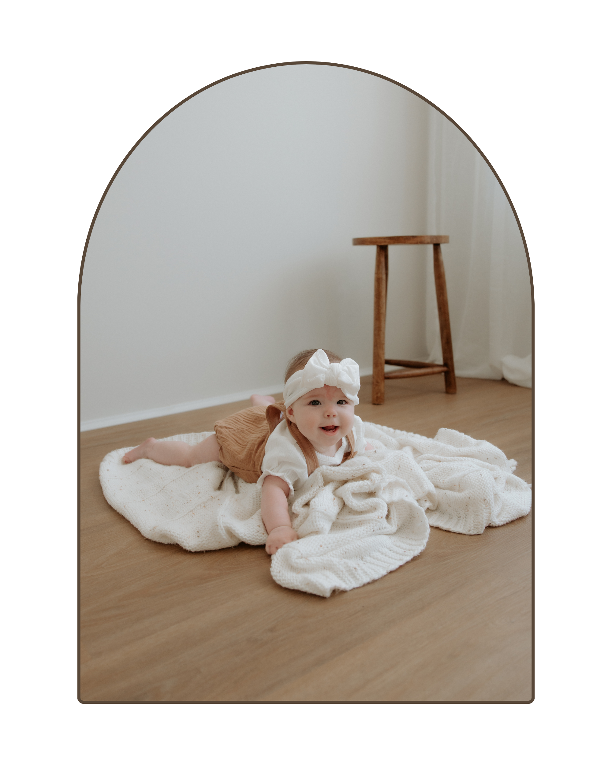 A baby girl with a white bow headband and tan shorts, lying on a cream-colored blanket on a wooden floor in front of a round mirror.