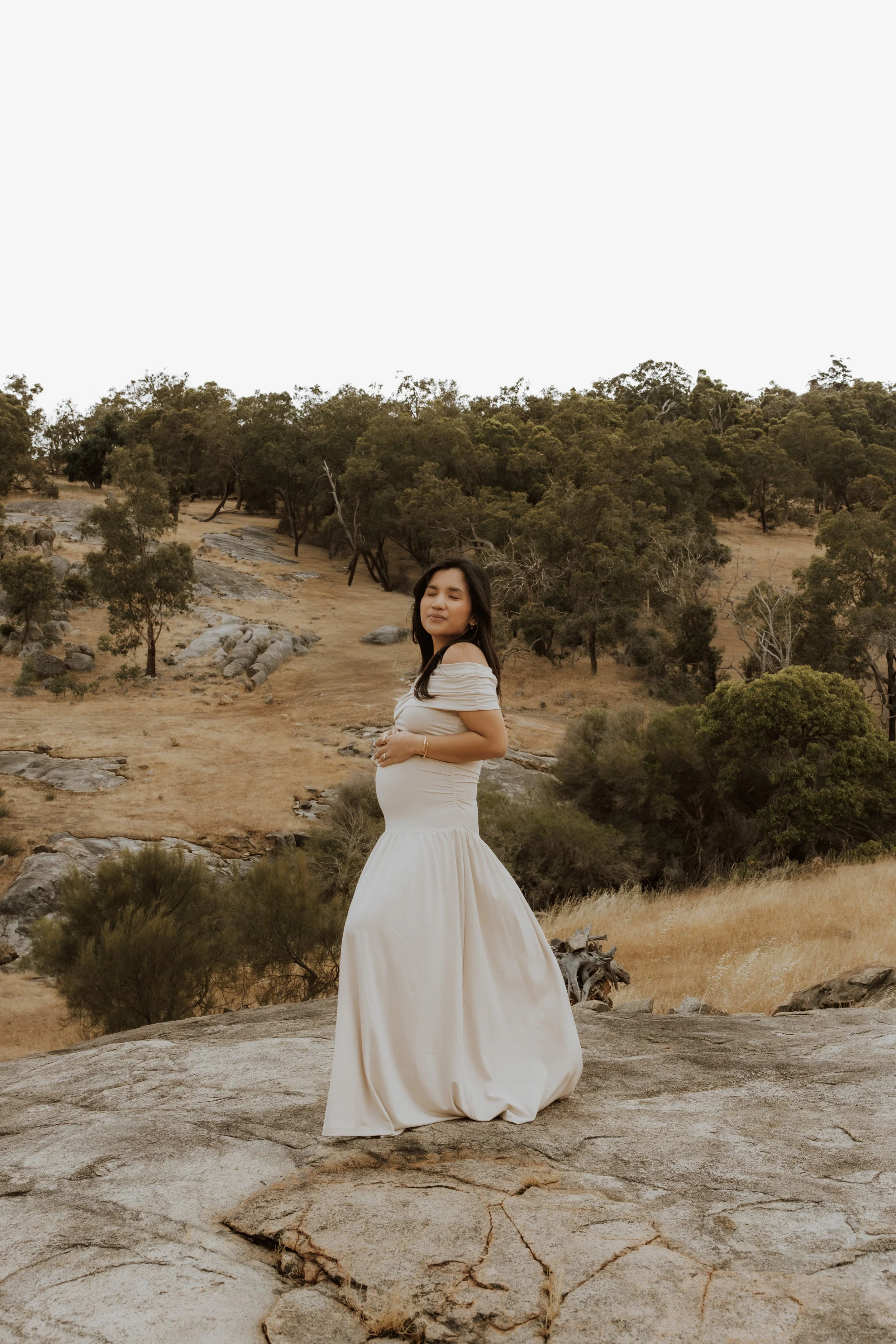 A pregnant woman in a white dress standing on a rocky surface in a scenic outdoor landscape with hills, trees, and dry grass in the Perth hills photoshoot