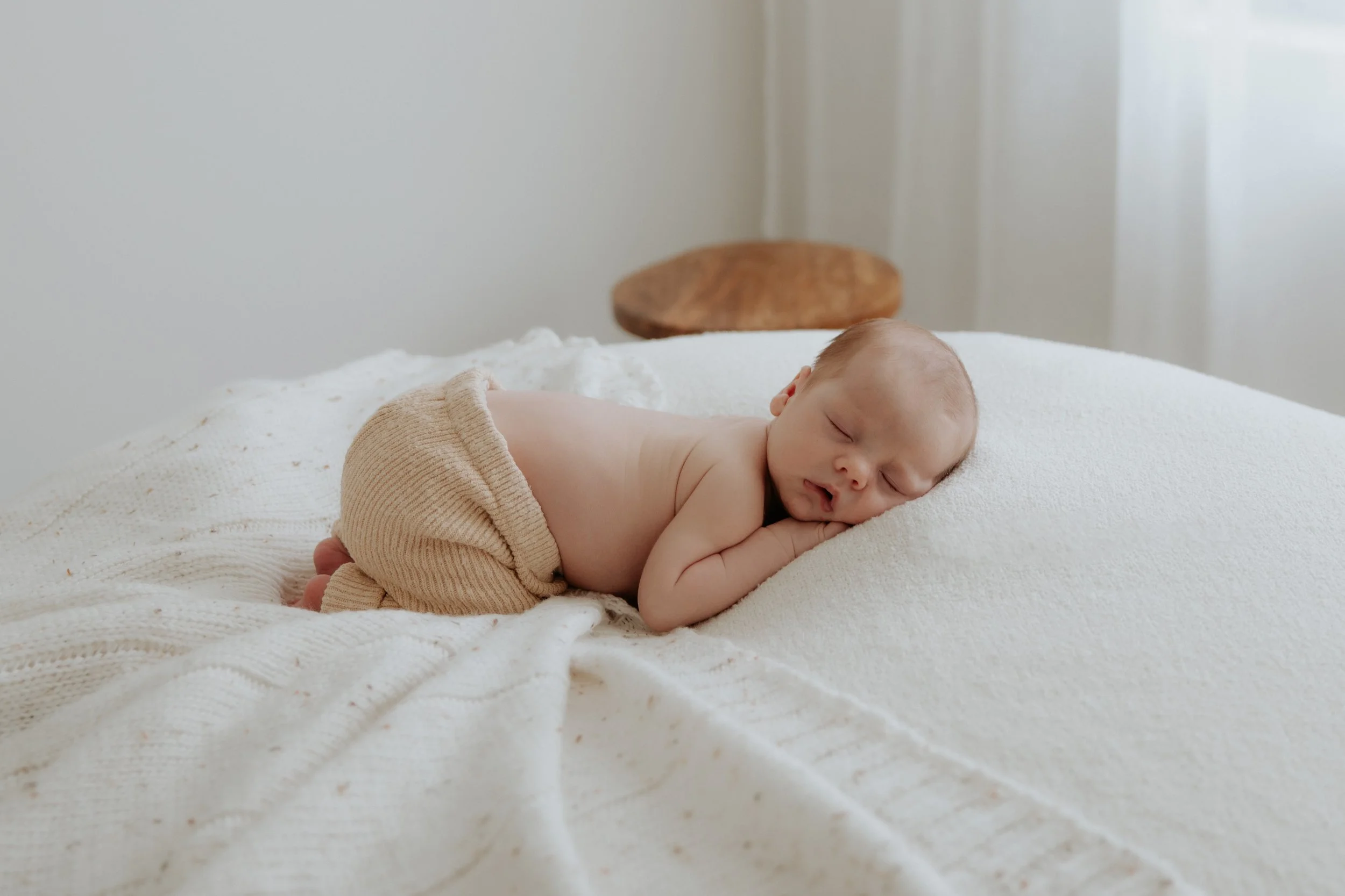 A sleeping baby laying on his side on a bed with white bedding, wearing beige knit shorts with a textured pattern. Simple, minimal but cozy studio newborn set up.