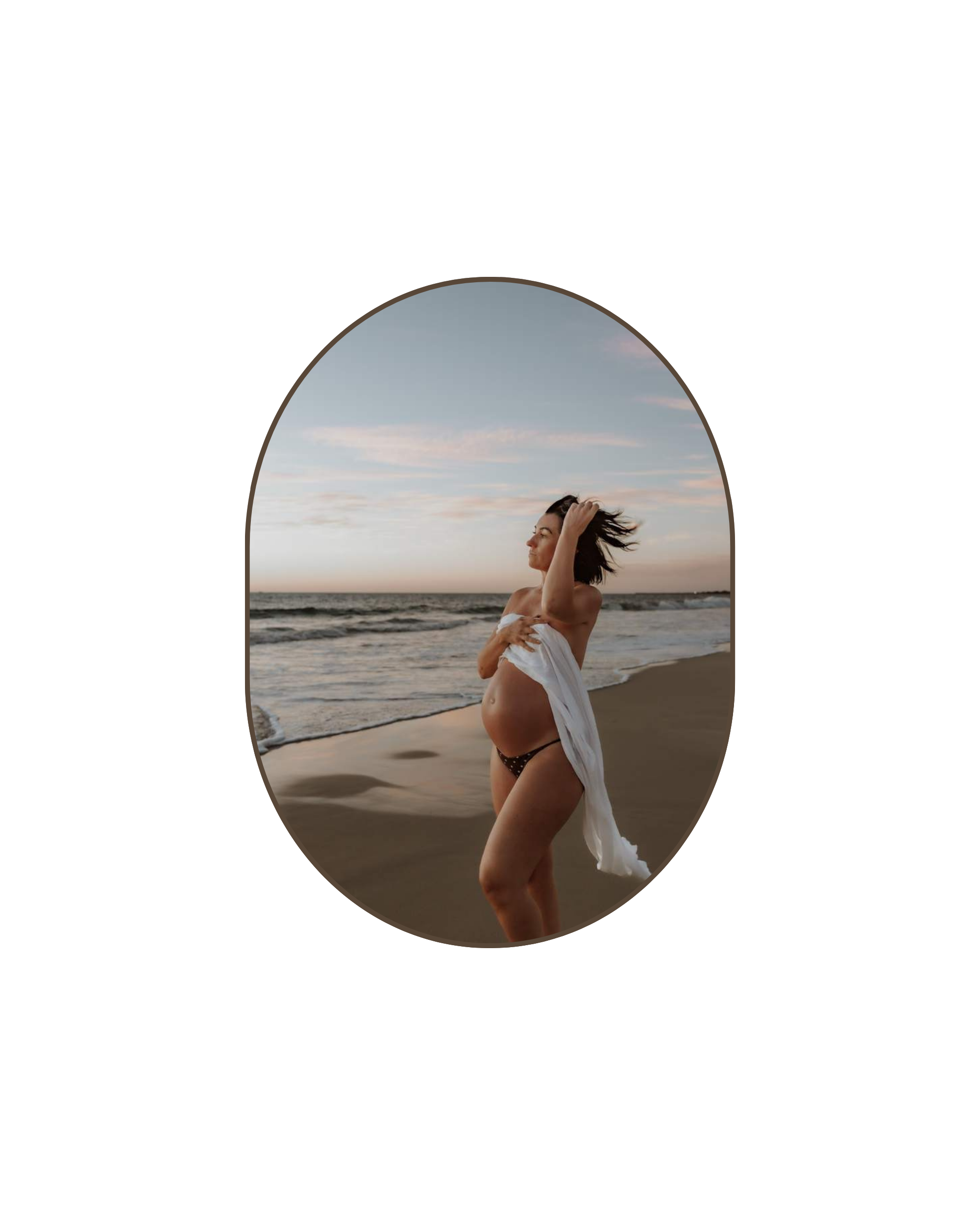 A pregnant woman standing on a beach at sunset, partially wrapped in a white cloth, with her hand on her head, looking out towards the ocean.