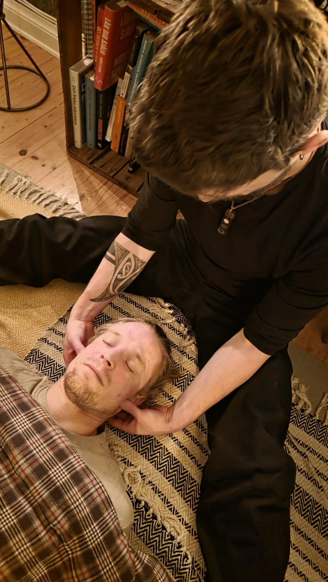 A person lying down with closed eyes on a patterned blanket, receiving a neck massage from another person. The person giving the massage has a tattoo on their arm and is wearing a black top. The scene is in a cozy room with a wooden floor and bookshelves in the background.