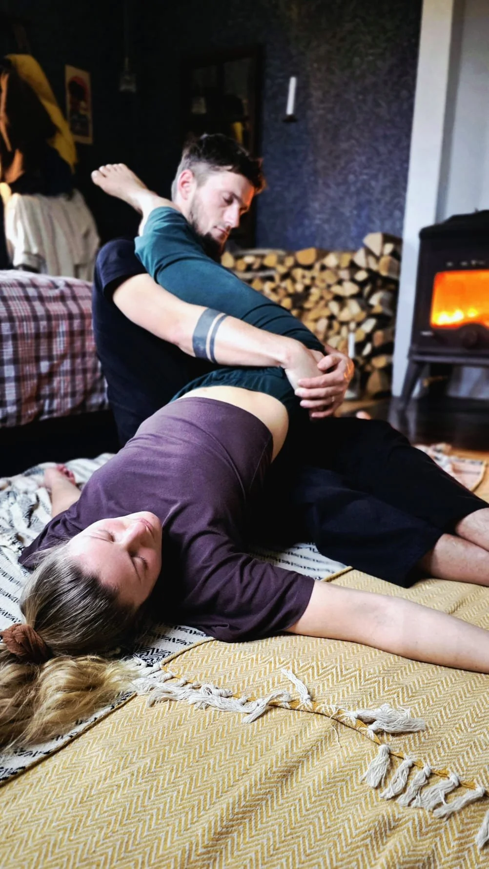 A man giving a massage to a woman lying on a rug in a cozy room with a fireplace and stacked firewood in the background.