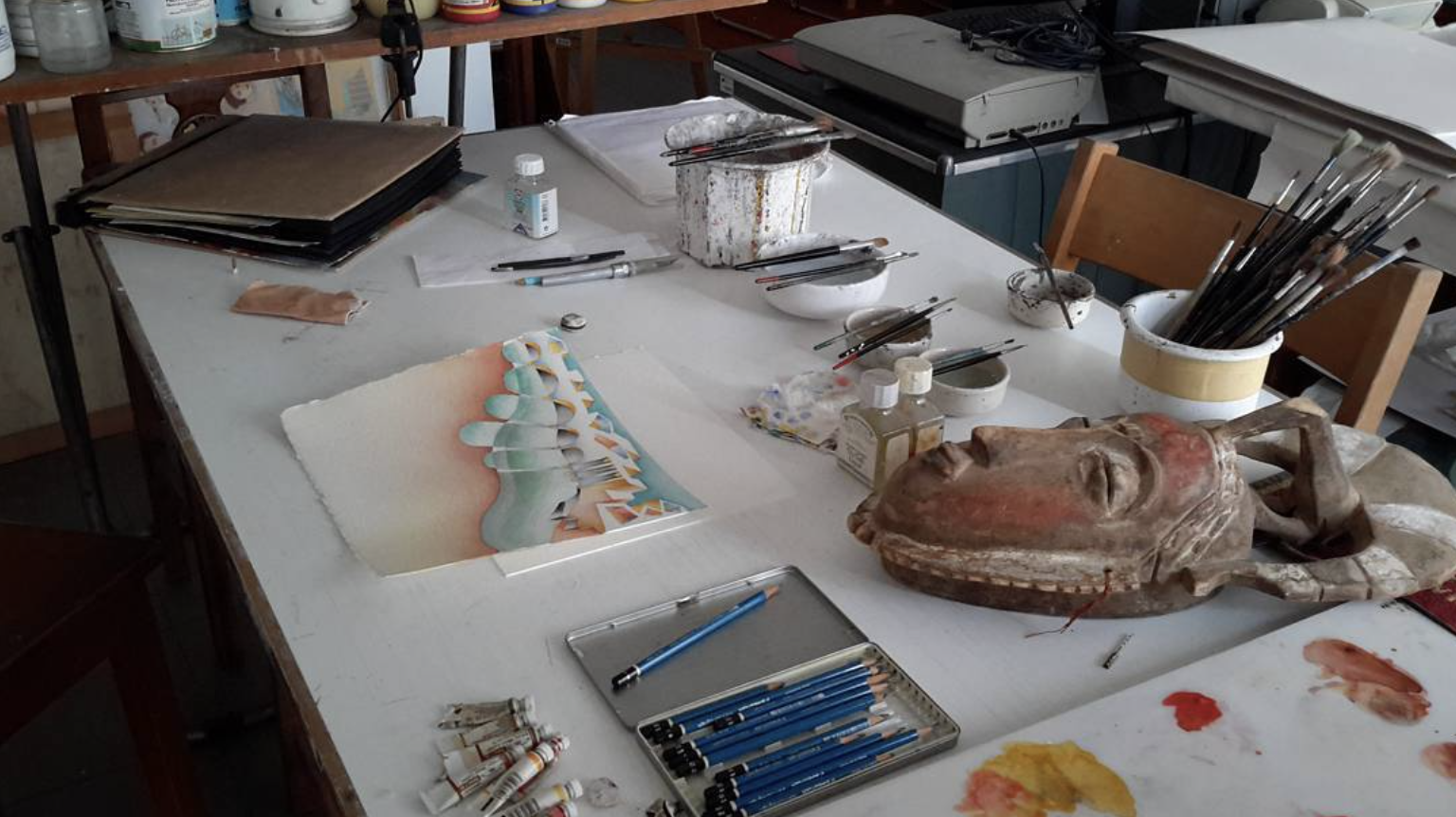 Mesa de estúdio de arte com pincéis, lápis, tubos de tinta, uma escultura, papéis e potes de tinta.