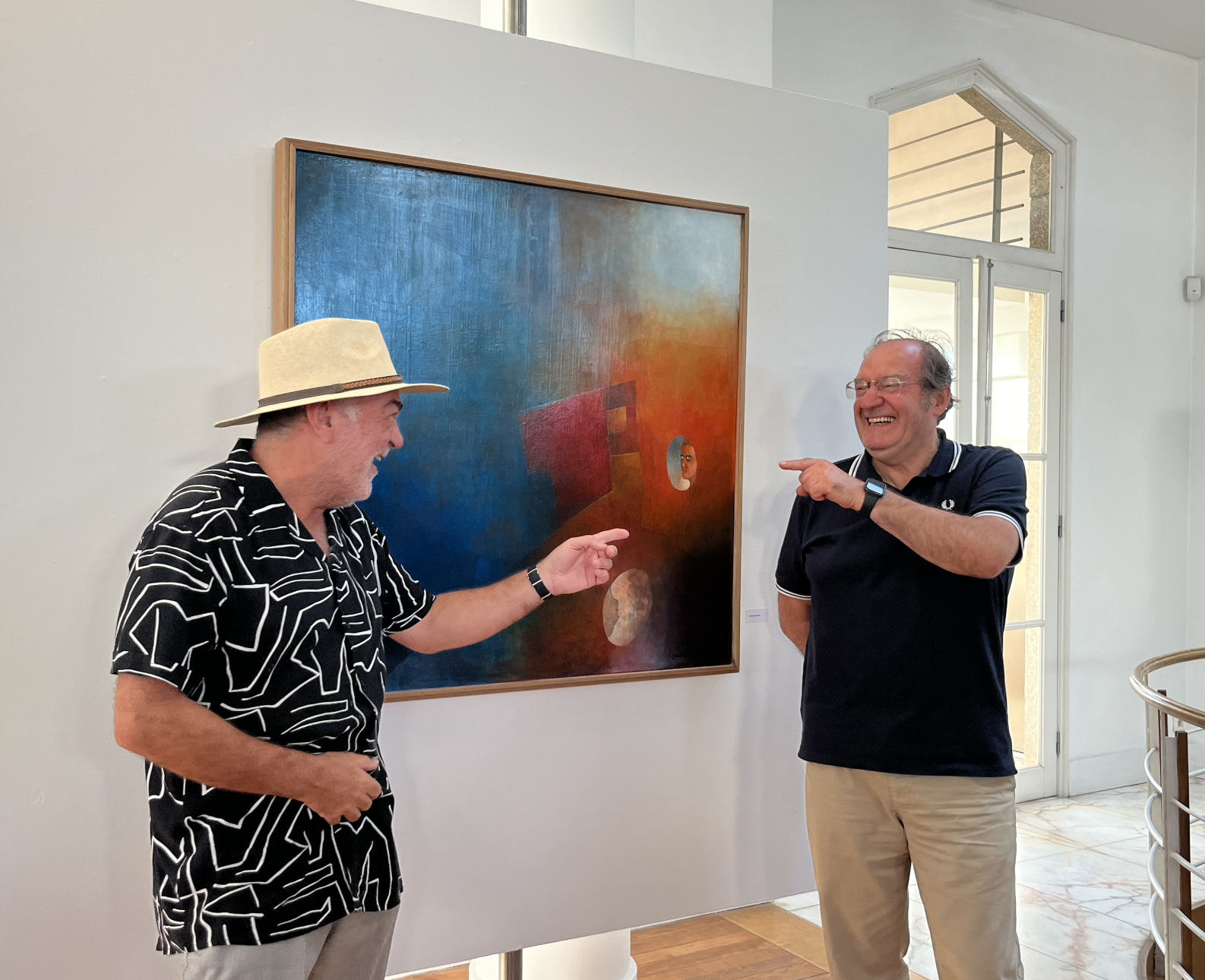 Dois homens sorrindo e apontando um para o outro em frente a uma pintura abstrata em uma galeria de arte.