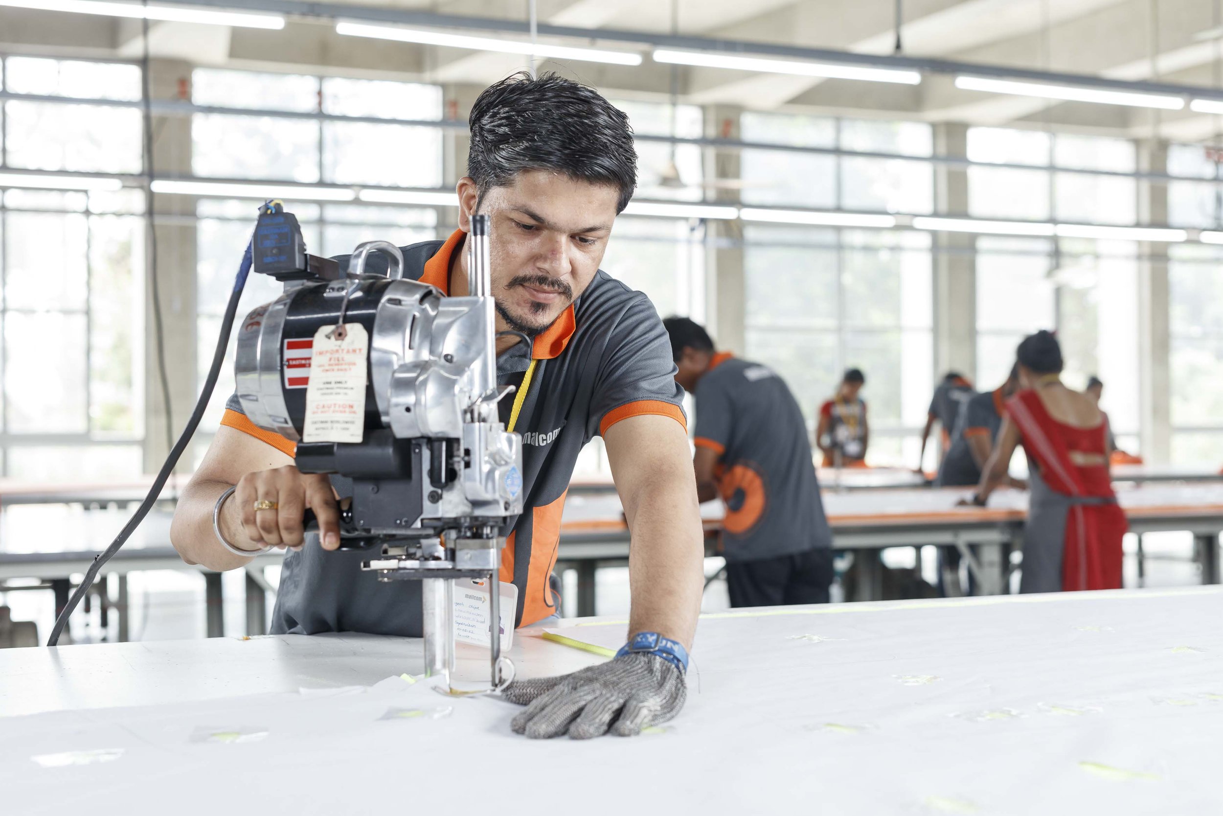 Man operating a sewing machine in a factory with large windows and other workers in the background.