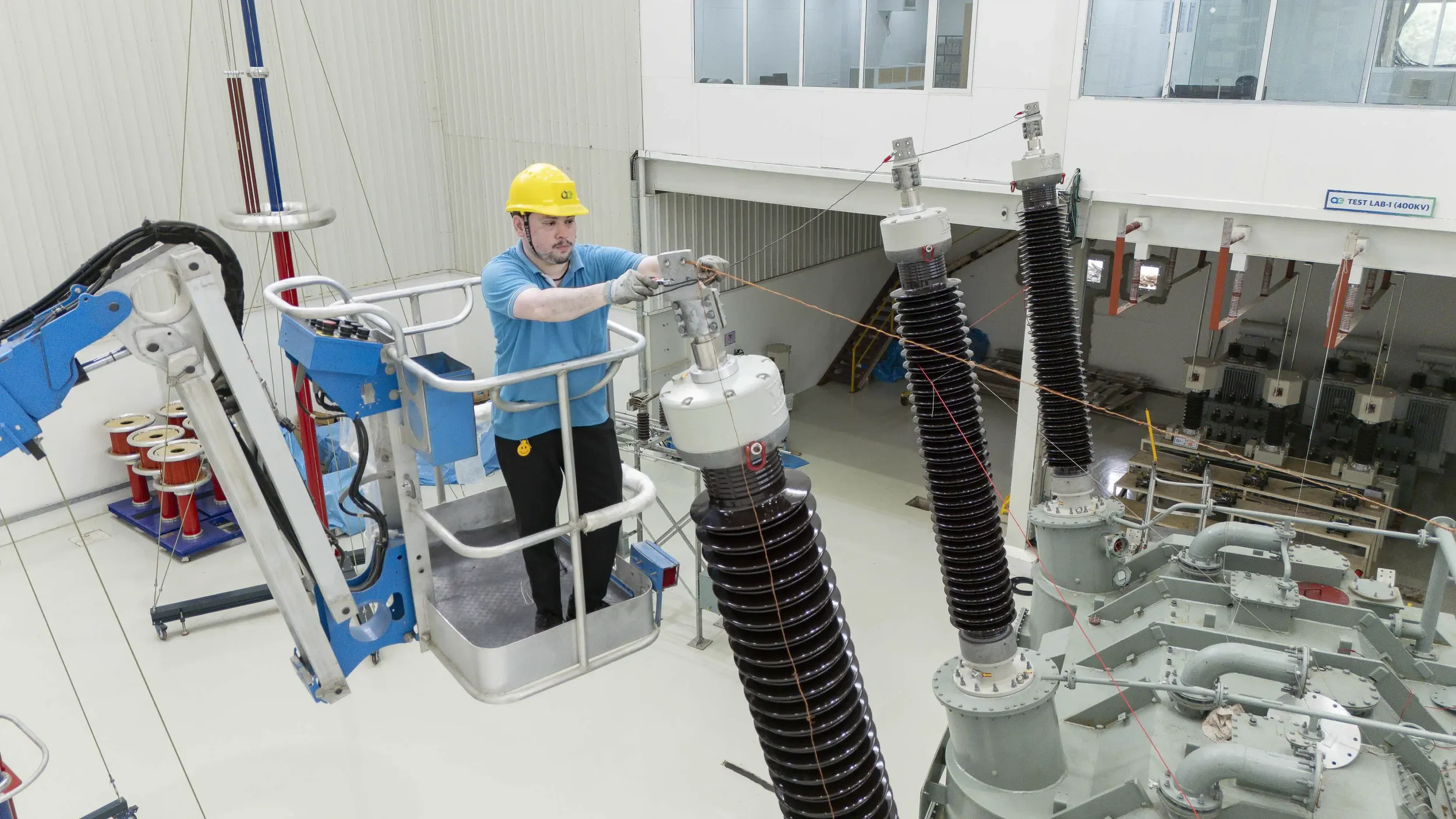 A technician in a safety harness and yellow helmet operating a lift to access high-voltage transformer bushings during testing.