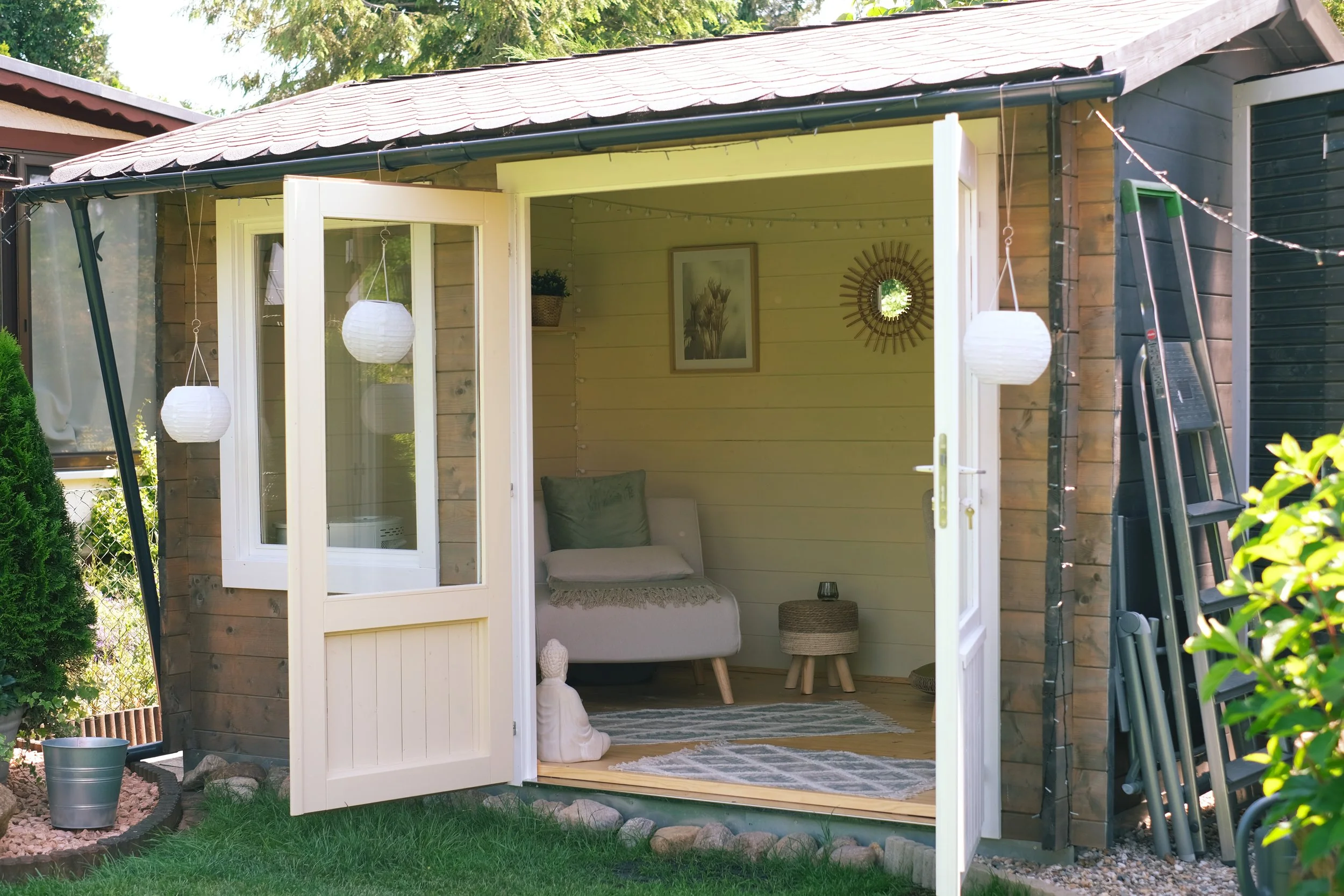 Ein offener Gartenpavillon mit einer Innenausstattung, inklusive einer weißen Couch, einem kleinen Beistelltisch, Wanddekorationen und Papierlaternen, im Garten mit grünem Rasen und Bäumen im Hintergrund.