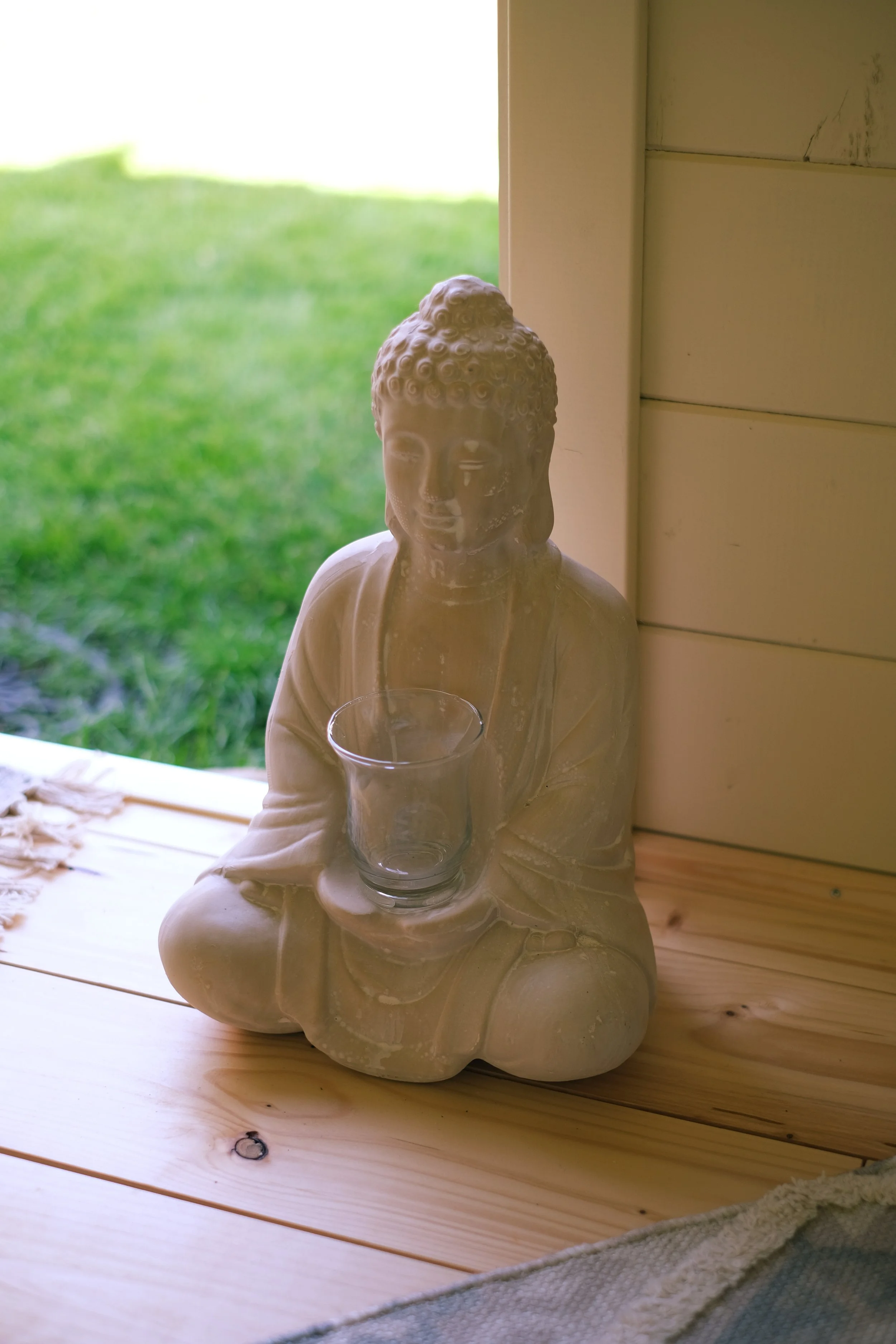 Steinfigur eines sitzenden Buddha mit einem Glas in der Hand, auf einem Holztisch neben einer Fensteröffnung mit Blick ins Grüne.