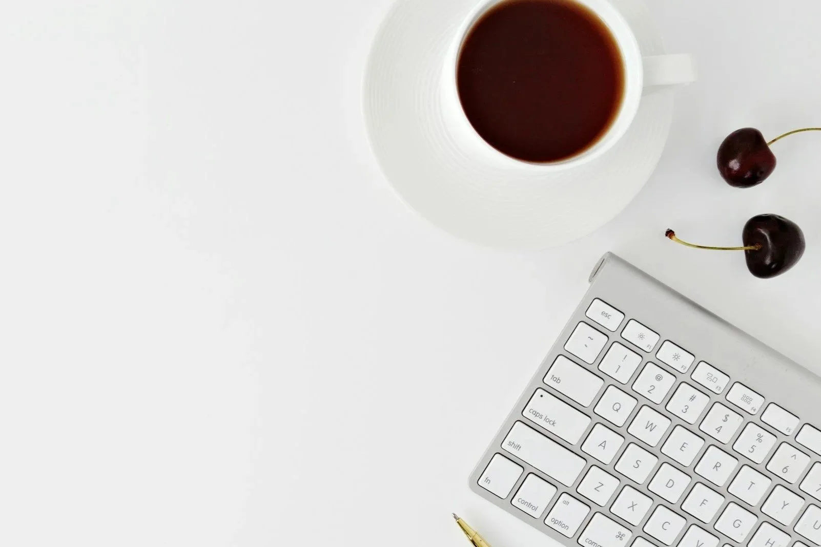 Top view of a white keyboard, a white coffee mug filled with coffee, two cherries, and a gold pen on a white surface.