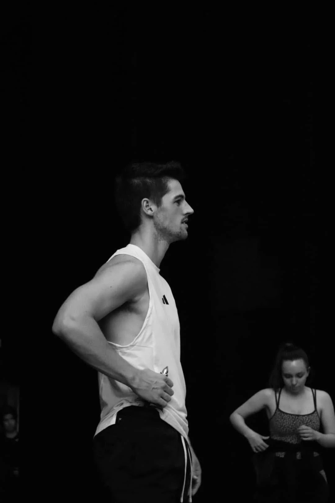 A young man with short dark hair, a trimmed beard, wearing a sleeveless athletic shirt, is standing in profile against a dark background. A young woman with long hair, wearing a tank top, is in the background looking down.