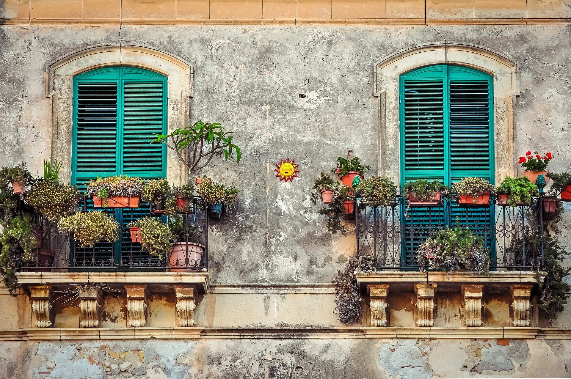 Two balconies with turquoise shutters and flower pots with plants, a small tree, and a sun decoration on a weathered wall.
