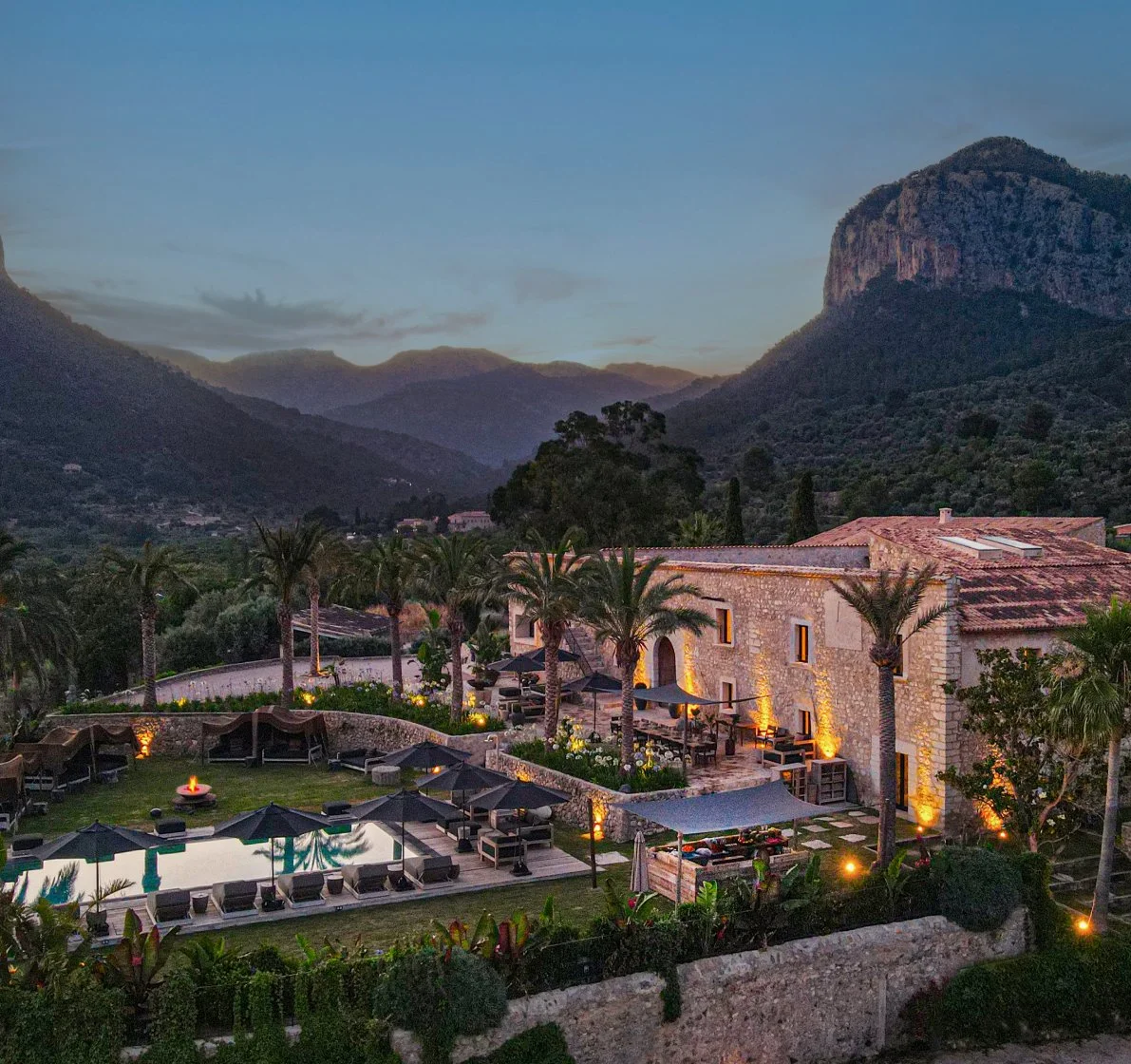 Luxury resort with swimming pool, palm trees, outdoor seating, and mountains in the background at dusk.