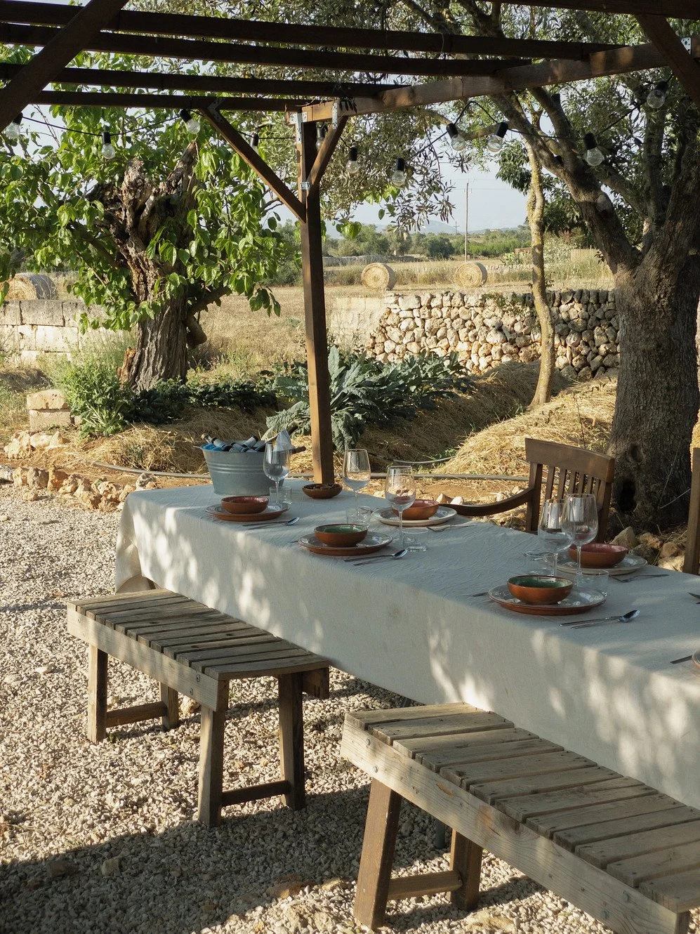 An outdoor dining table set for a meal under a wooden pergola with string lights. The table has a white tablecloth, bowls, glasses, and cutlery. There are trees, a stone wall, and hay bales in the background.