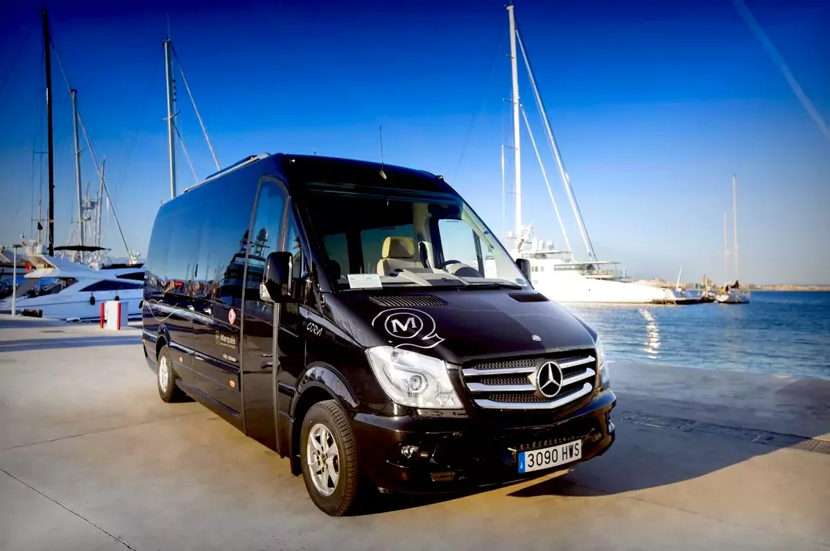 Black Mercedes-Benz van parked near a marina with sailboats and yachts in the background, clear blue sky, and calm water.