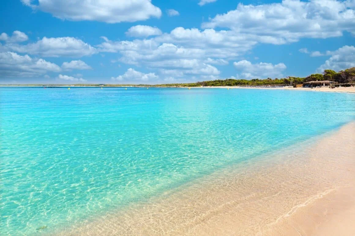 A tropical beach with clear turquoise water, soft sandy shoreline, and a partly cloudy blue sky.