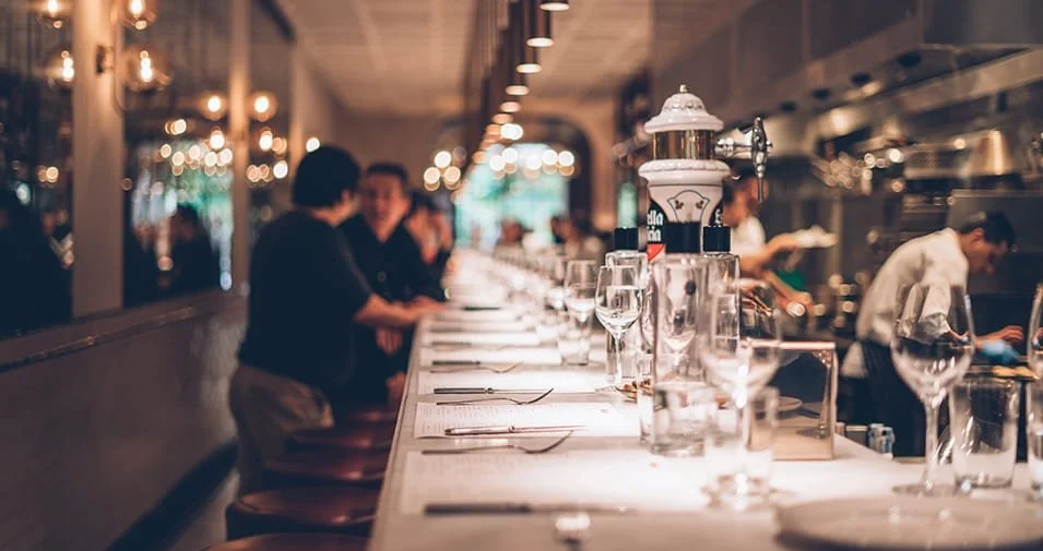A bar with a long counter set with wine glasses, plates, and utensils, in a dimly lit restaurant with small lights and people dining and chatting.