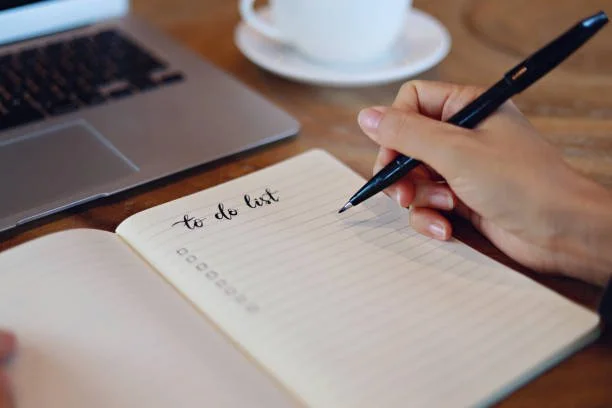 A person writing 'to do list' in a notebook with a black pen, a laptop, and a cup of coffee on a wooden table.
